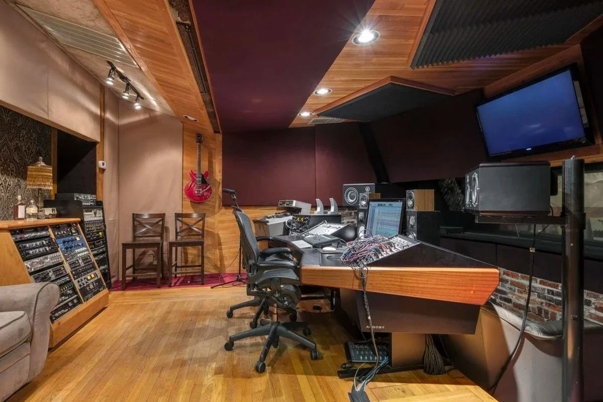 Recording studio control room with mixing console, studio monitors, computer, TV monitor, guitars on wall, chairs, and acoustic panels.