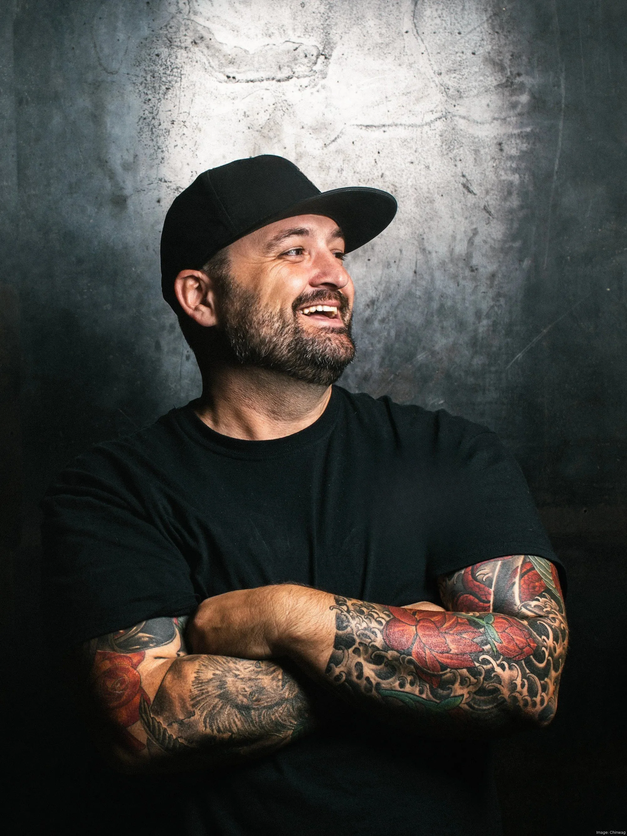A smiling man with a beard and tattooed arm wearing a white shirt and black apron, standing in a commercial kitchen.