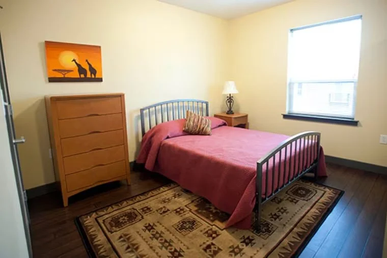 A bedroom with a bed, a dresser, a bedside table with a lamp, and a window with sunlight, featuring a patterned rug on hardwood floor.
