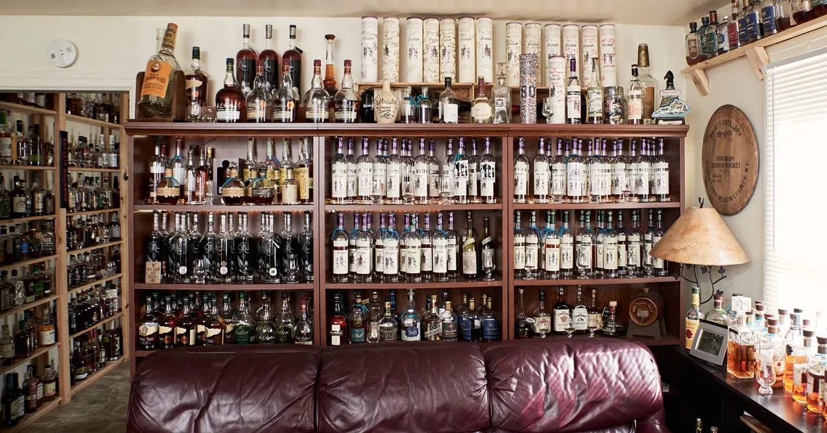 A room with wooden shelves filled with numerous bottles of alcohol, including whiskey and wine. There is a burgundy leather sofa in front of the shelves, a lamp on the right side, and a window with blinds.