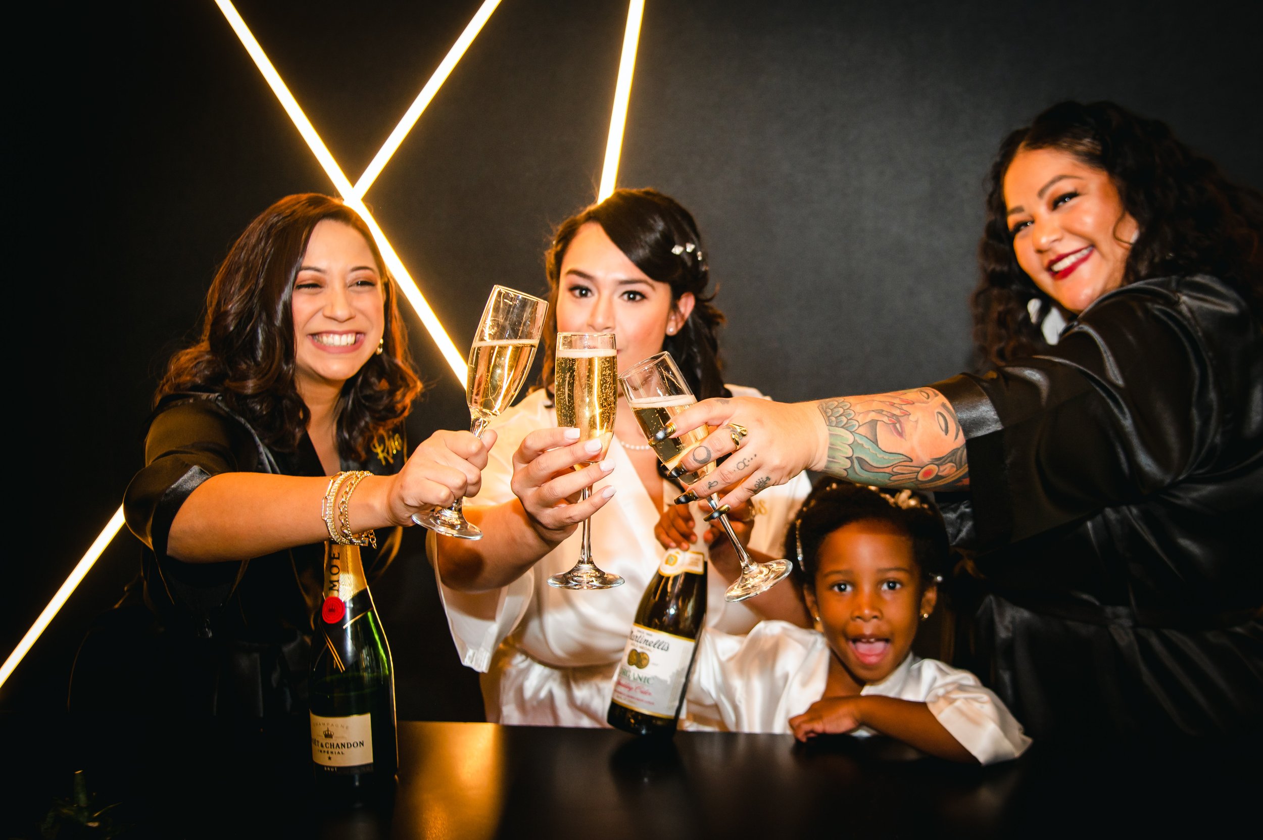 Group of women and a young girl celebrating with champagne glasses raised in a toast, with a dark background and neon lights.