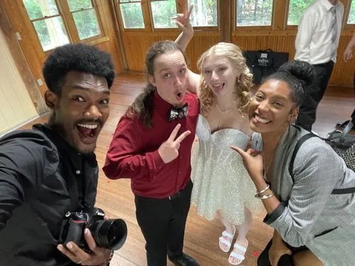 Four young people taking a selfie, smiling and making fun expressions, inside a wooden-paneled room with large windows, dressed in semi-formal attire.