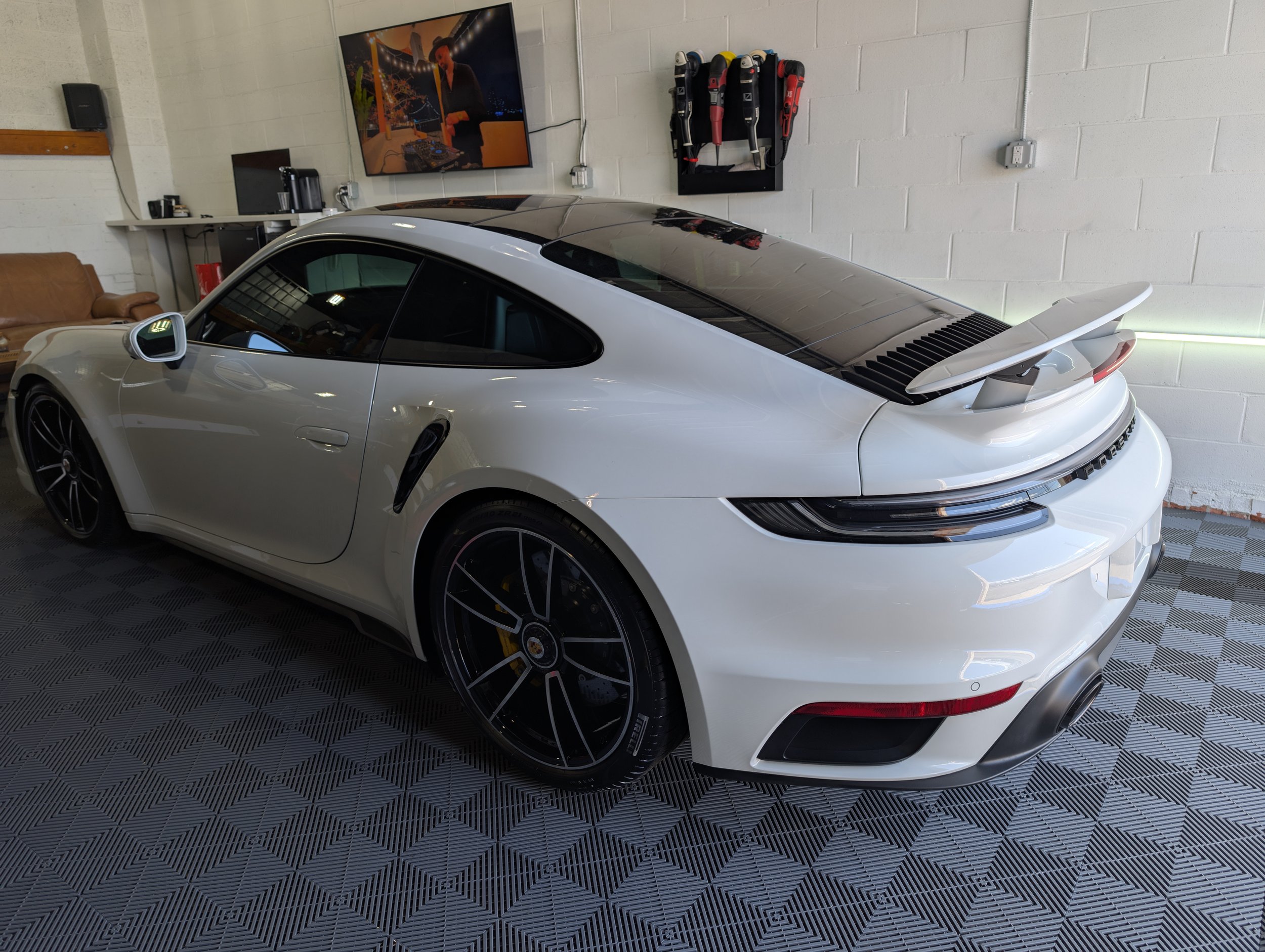 White sports car with a rear spoiler parked indoors on a tiled floor, with a television mounted on the wall showing a person, and tools hanging nearby.
