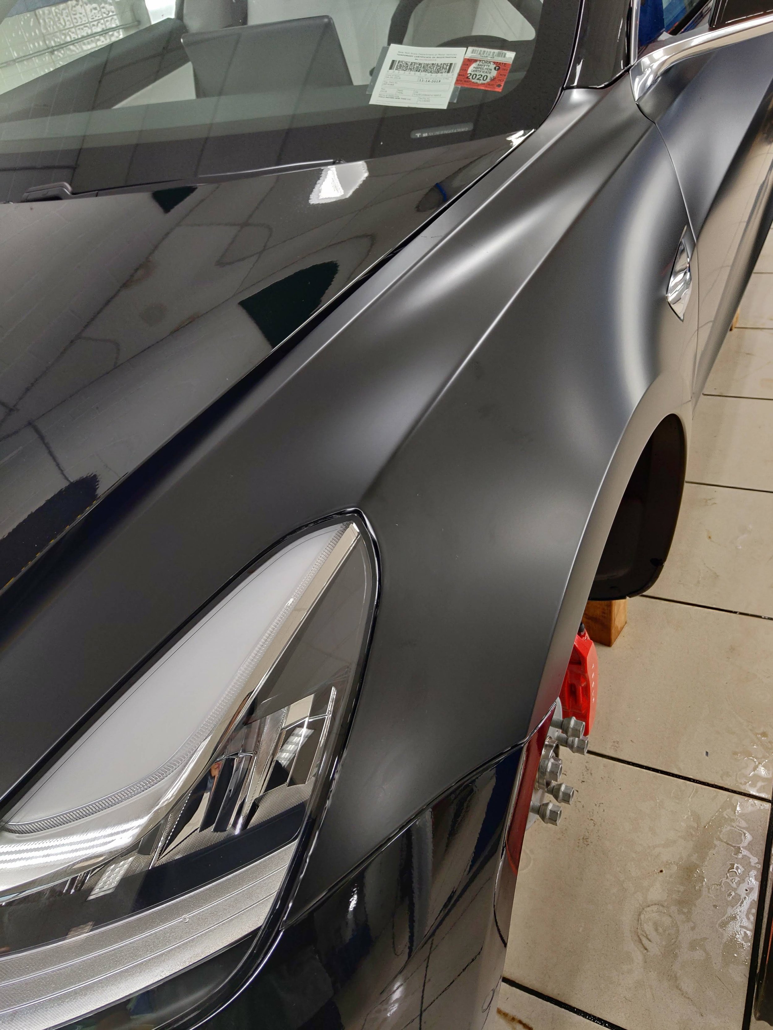 Close-up of a car's front-left side, showing a sleek matte black fender, headlight, and red brake caliper. The car is in a garage, possibly undergoing maintenance, with a part of the windshield and dashboard visible.