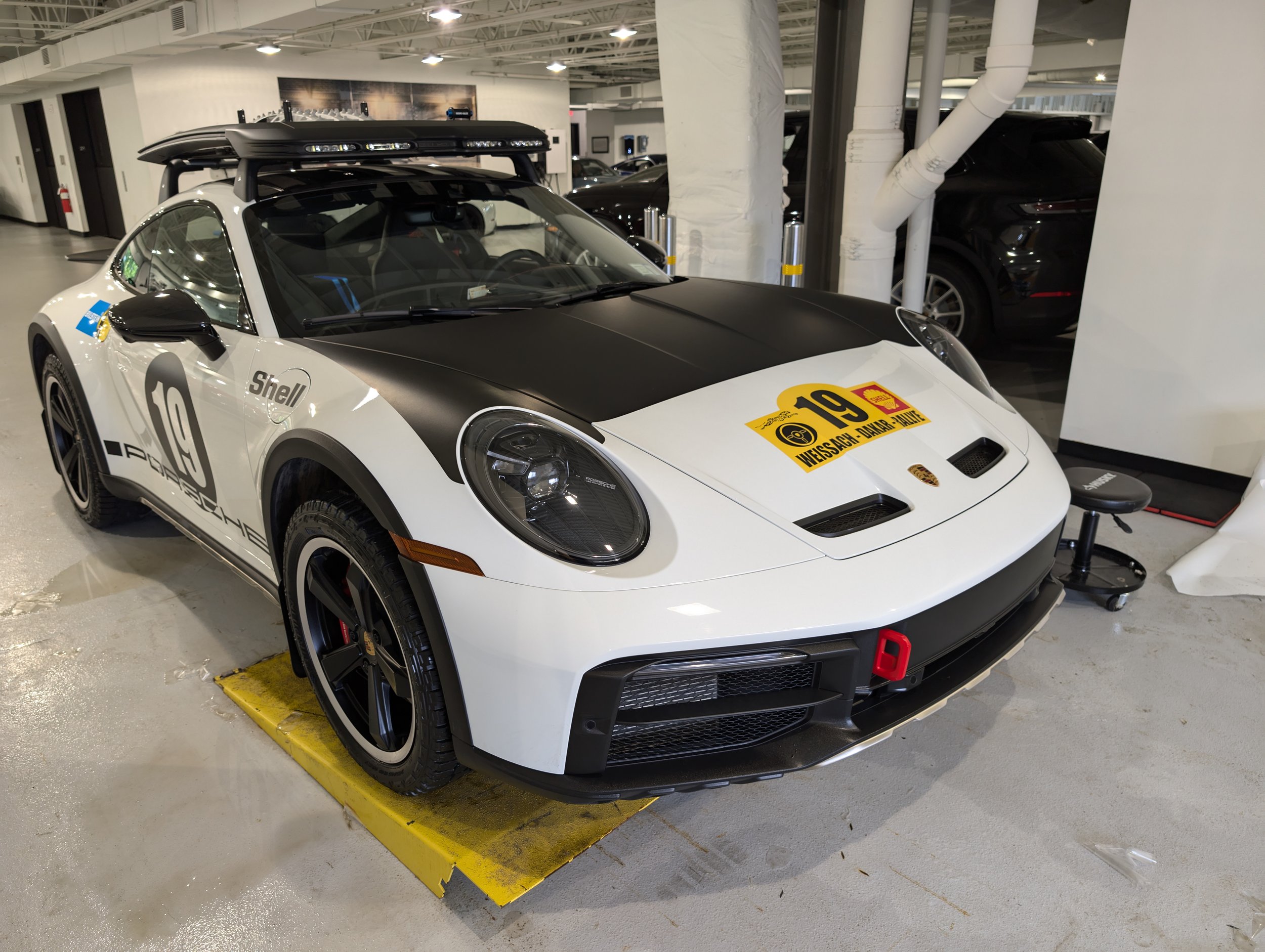 White Porsche sports car with black accents, race decals including the number 19, parked indoors, equipped with a roof rack, and sponsor logos from Shell.