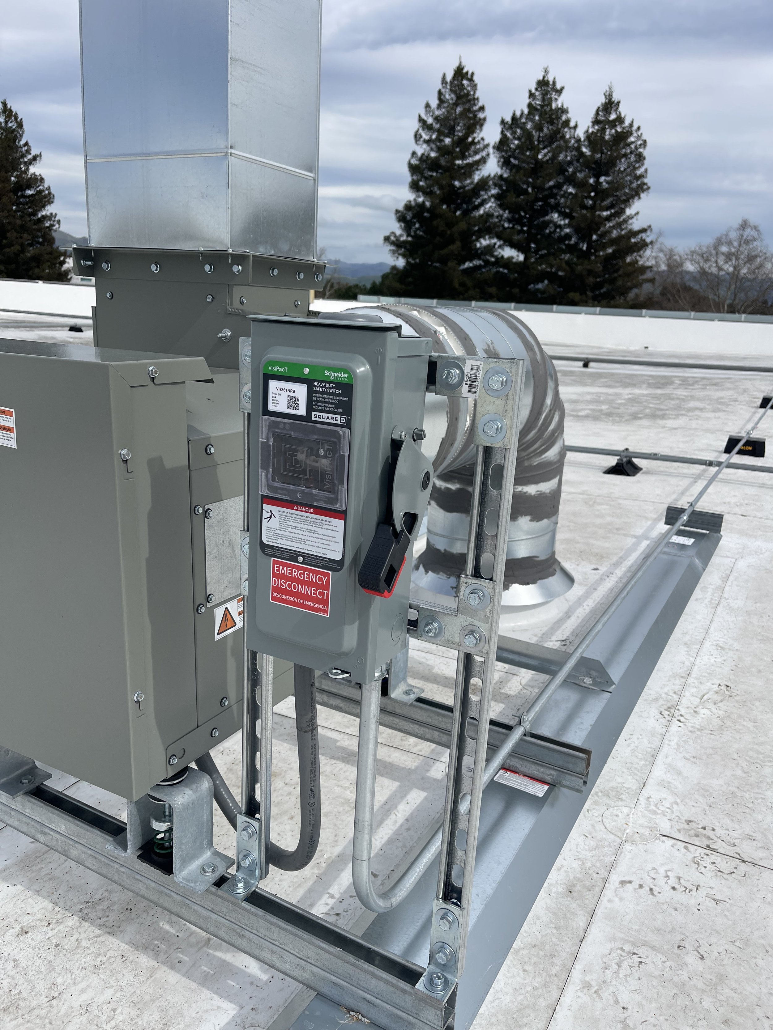 HVAC rooftop unit with electrical control box and ductwork, surrounded by a metal platform and some trees in the background.