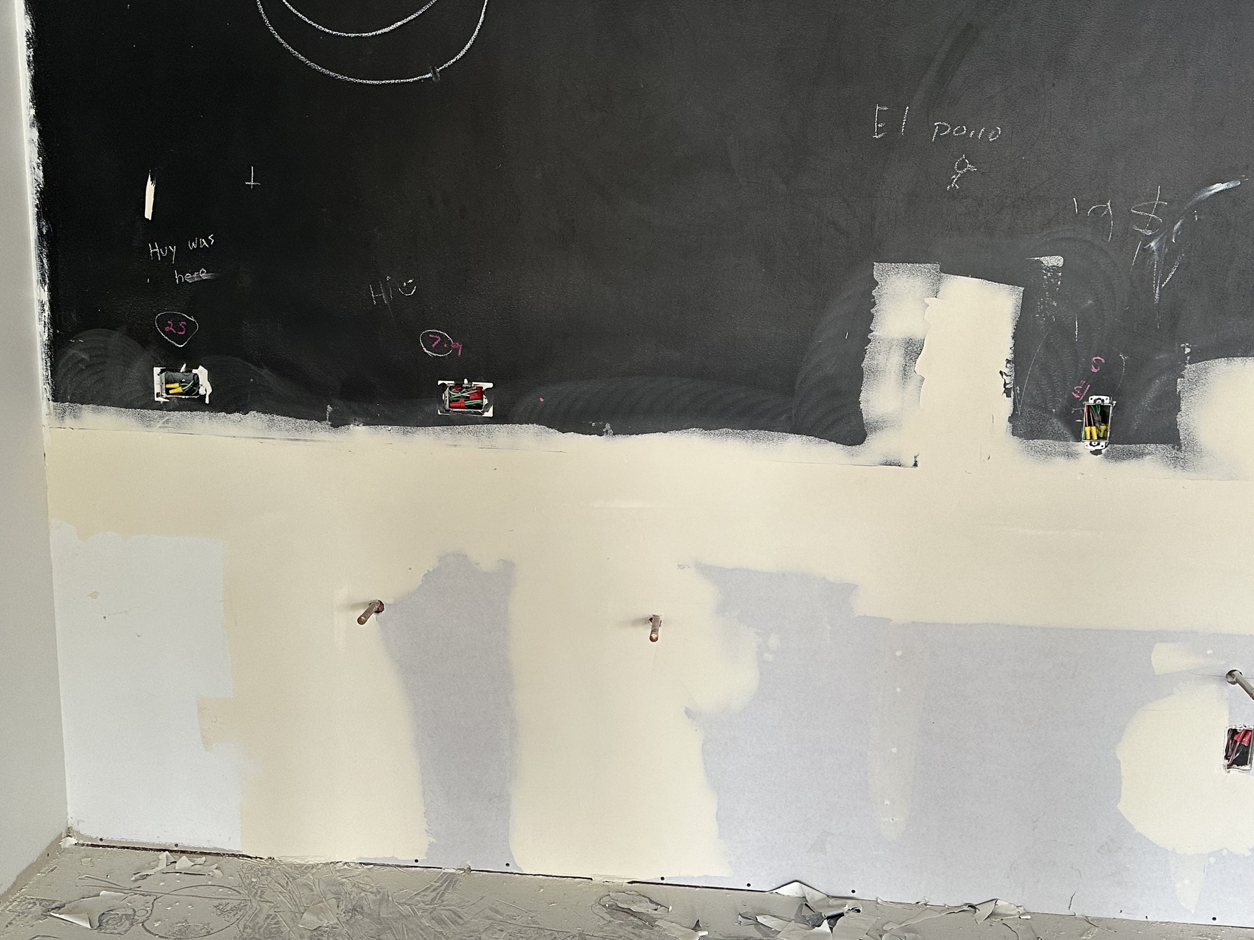 Partially painted wall with electrical outlets, handwritten notes, and small wiring boxes, some drywall patches visible.