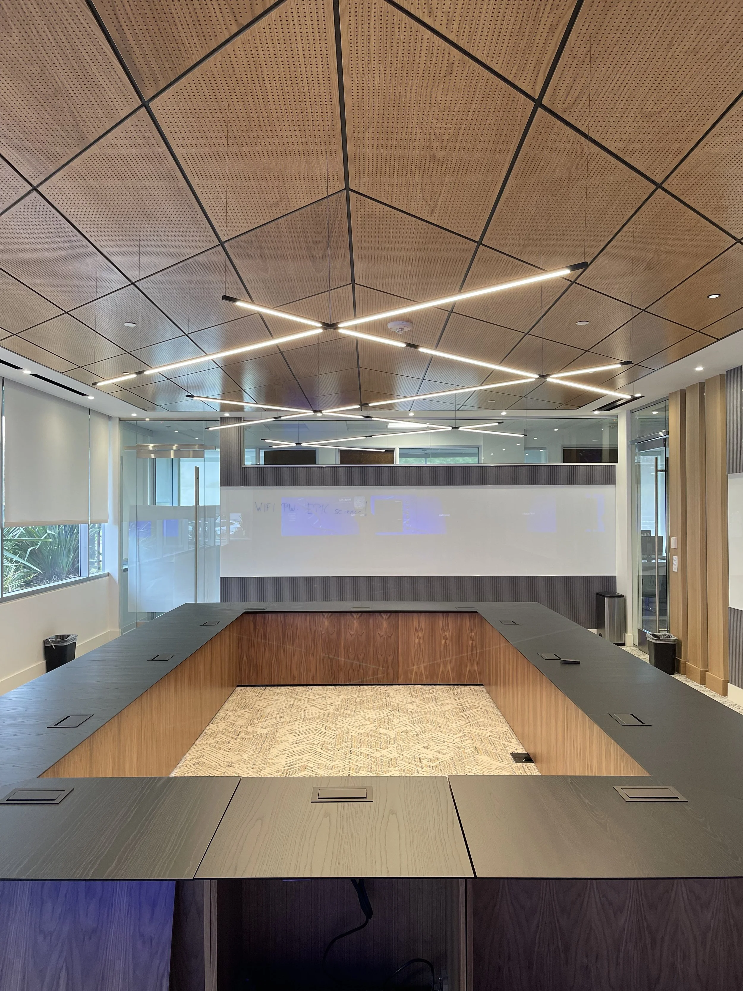 Modern conference room with a rectangular table, black tabletop surfaces, and a wooden interior at the center. The ceiling has a geometric design with LED lighting, and the room features large windows and glass walls, with some plants visible outside