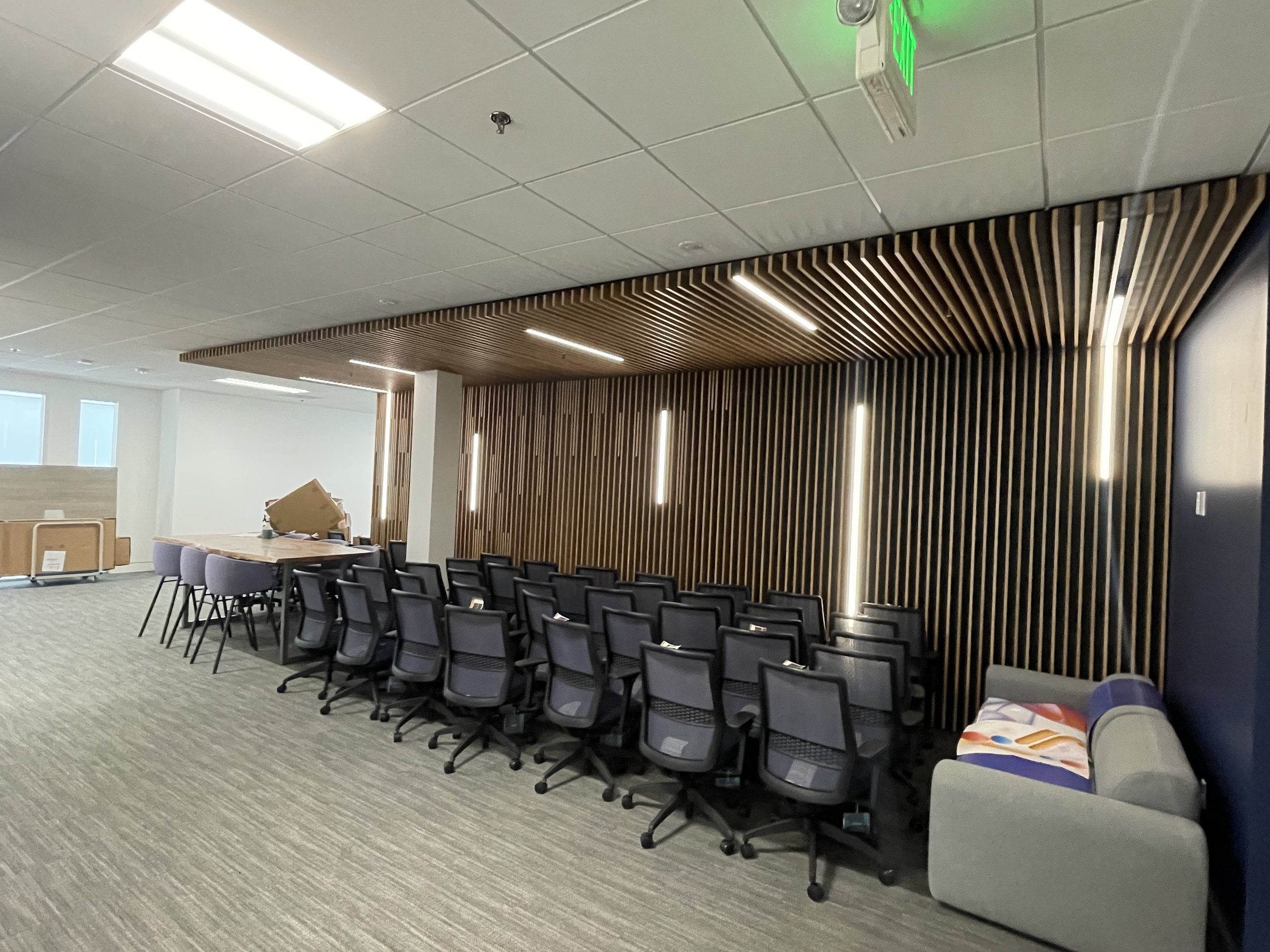 Empty conference room with black chairs arranged in rows facing a wooden slat wall with slit lights. A gray couch with a colorful magazine is to the right. To the left, there is a wooden table with chairs and some cardboard boxes, suggesting the spac