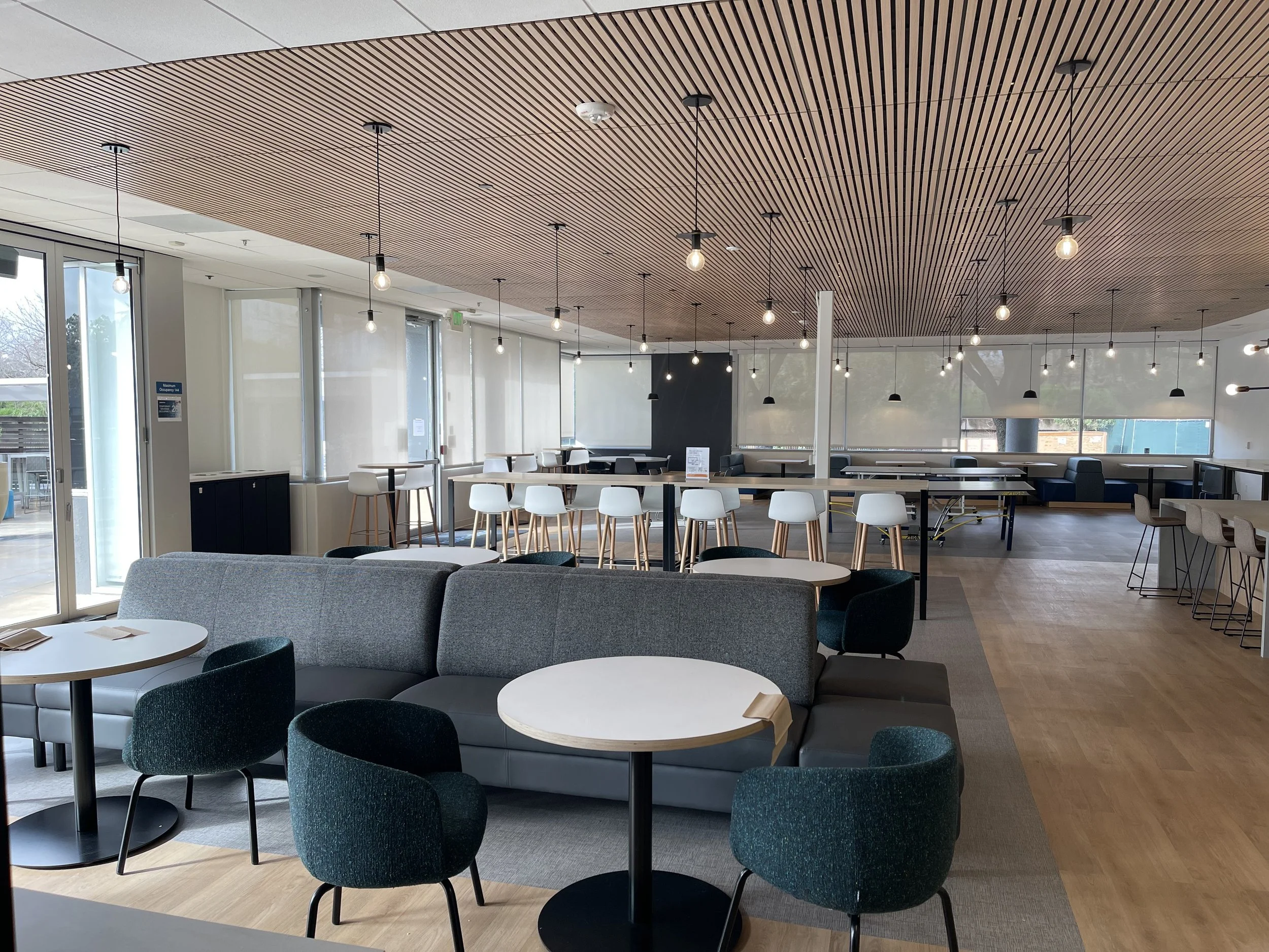 Modern, spacious lounge with round tables, chairs, and a sectional sofa, illuminated by hanging pendant lights and large windows.