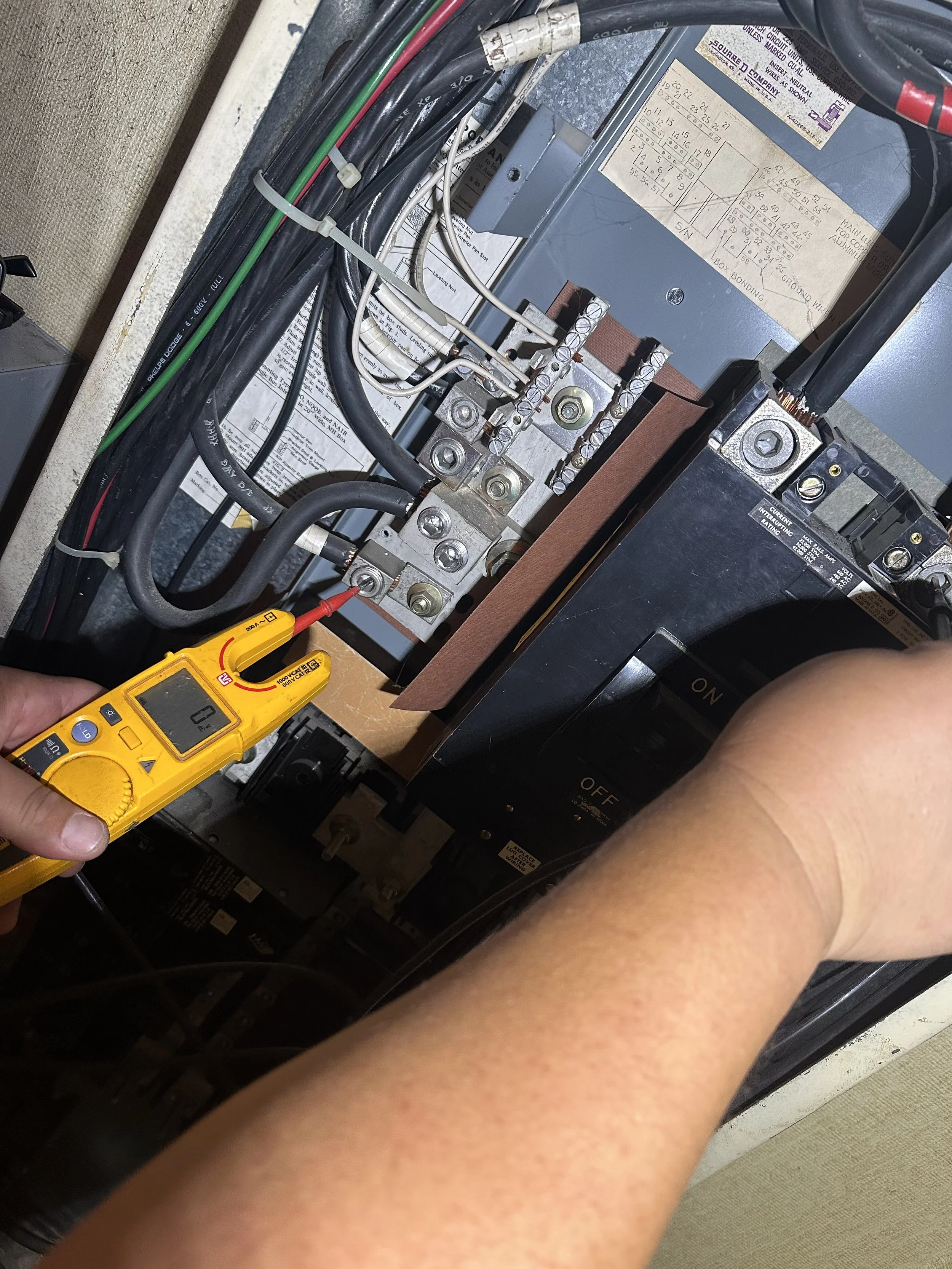 Close-up of an electrician using a digital multimeter to test electrical wires connected to an industrial control panel with circuit breakers and wiring.
