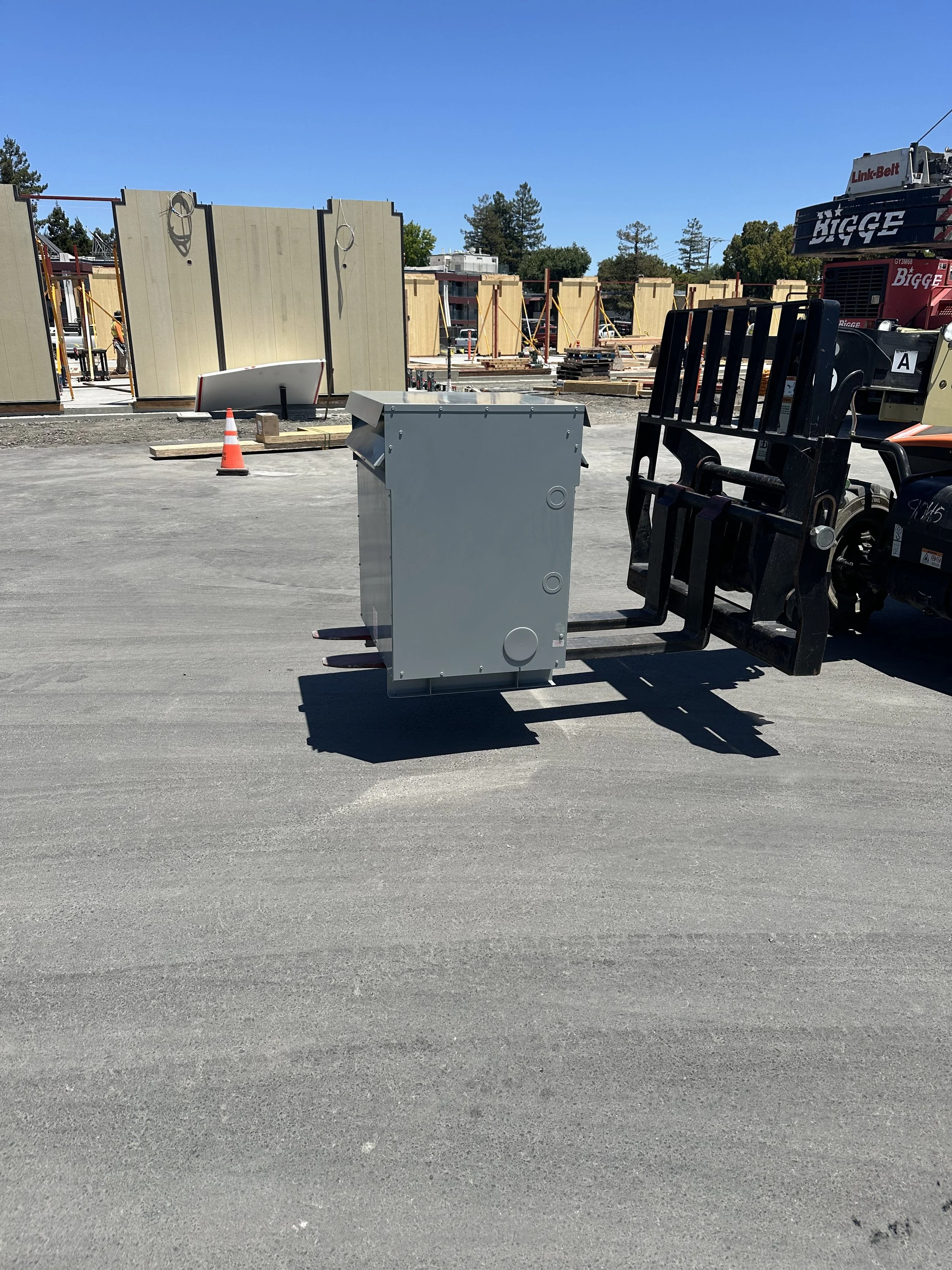 Construction site with building frames, a gray electrical box, and a forklift lifting the box.