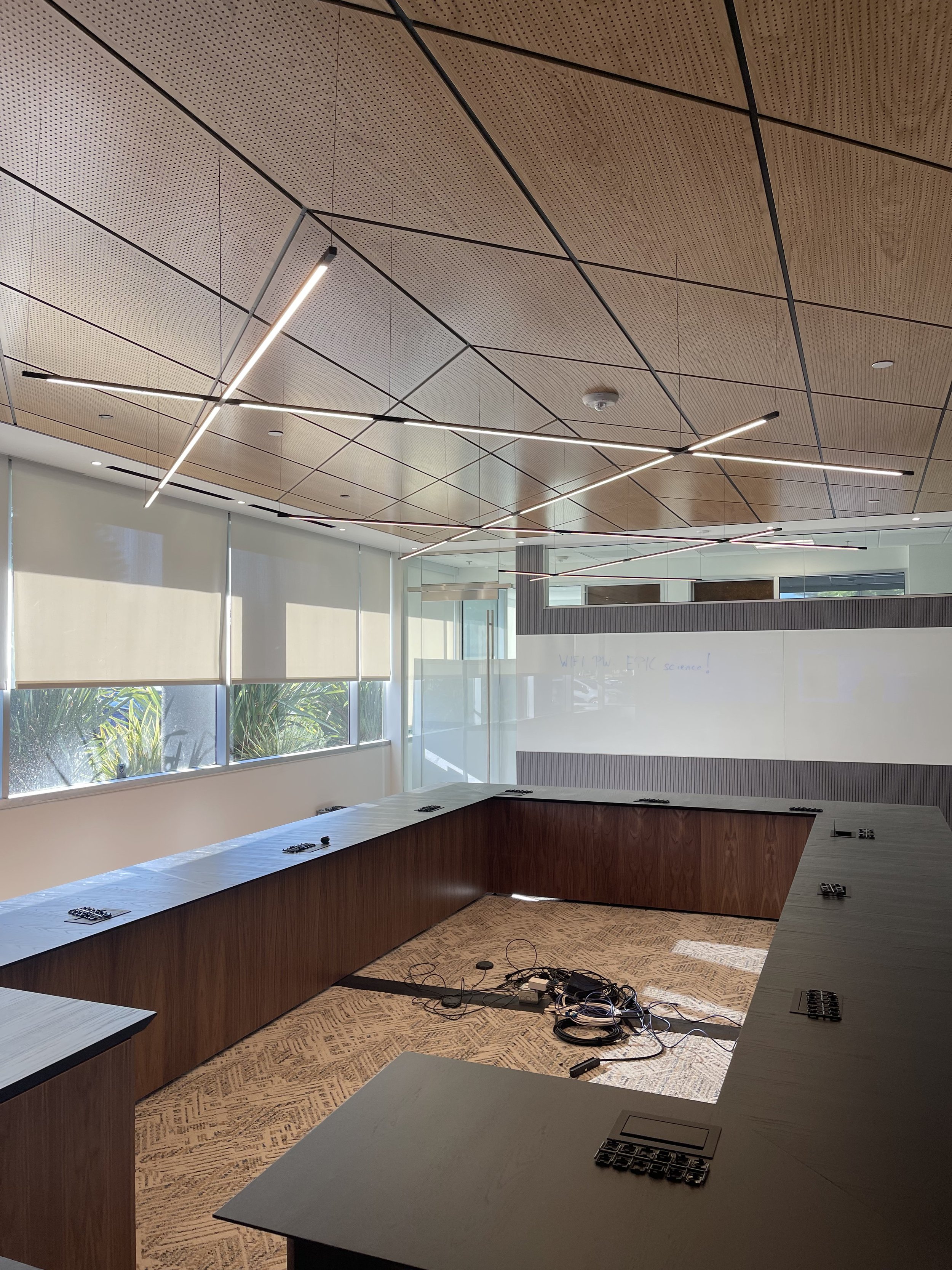 An empty modern conference room with wooden and gray tables arranged in a U-shape, large windows with blinds, whiteboard with faint writing, and a patterned carpet. There are wires and electronic equipment on the floor.