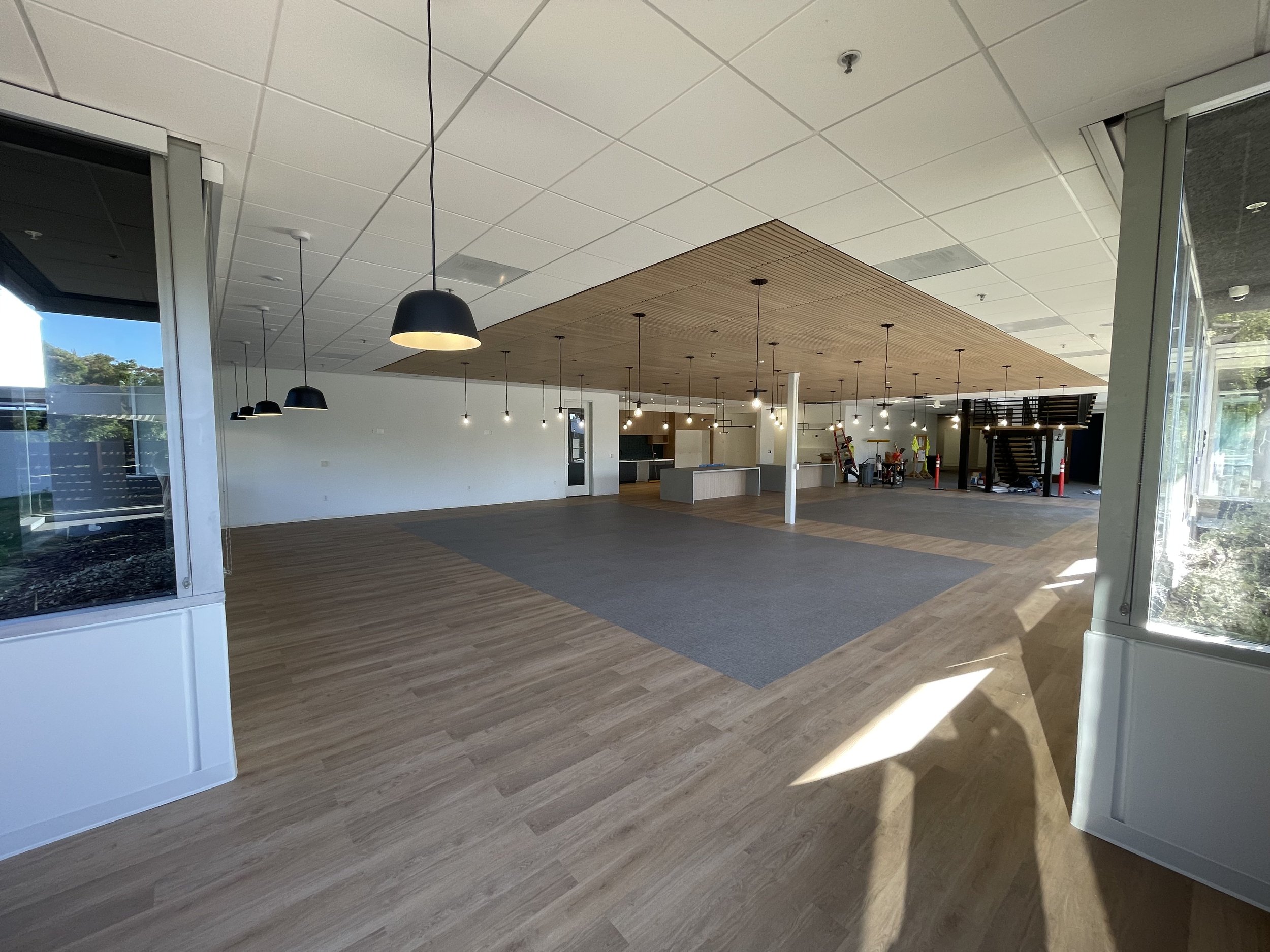 Empty interior space with wood flooring, white walls, and modern lighting fixtures hanging from the ceiling. There are large windows letting in natural light, and a kitchen area with a central island in the background.