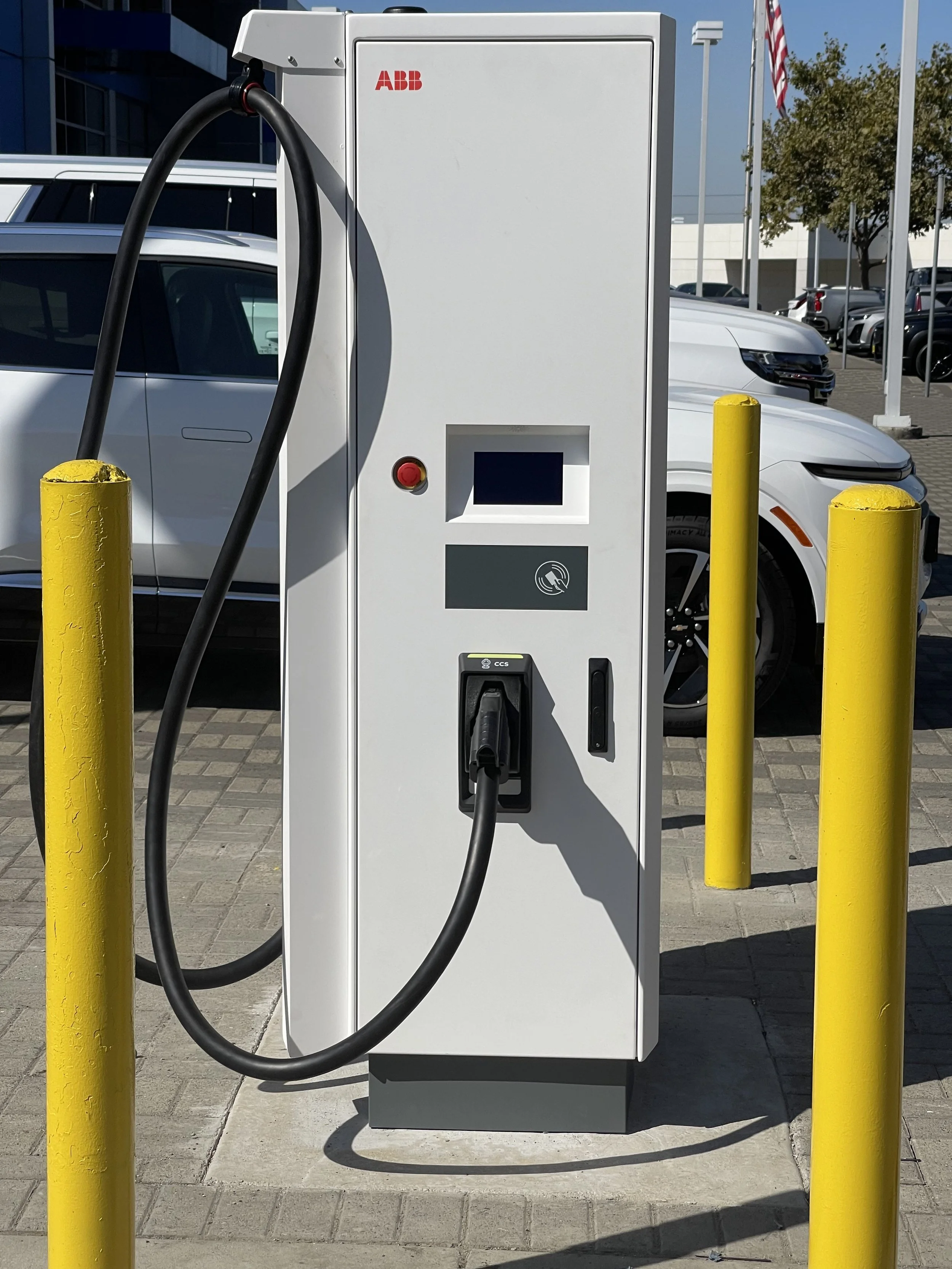 Electric vehicle charging station with a plugged-in cable, located on a paved parking lot with cars and trees in the background.