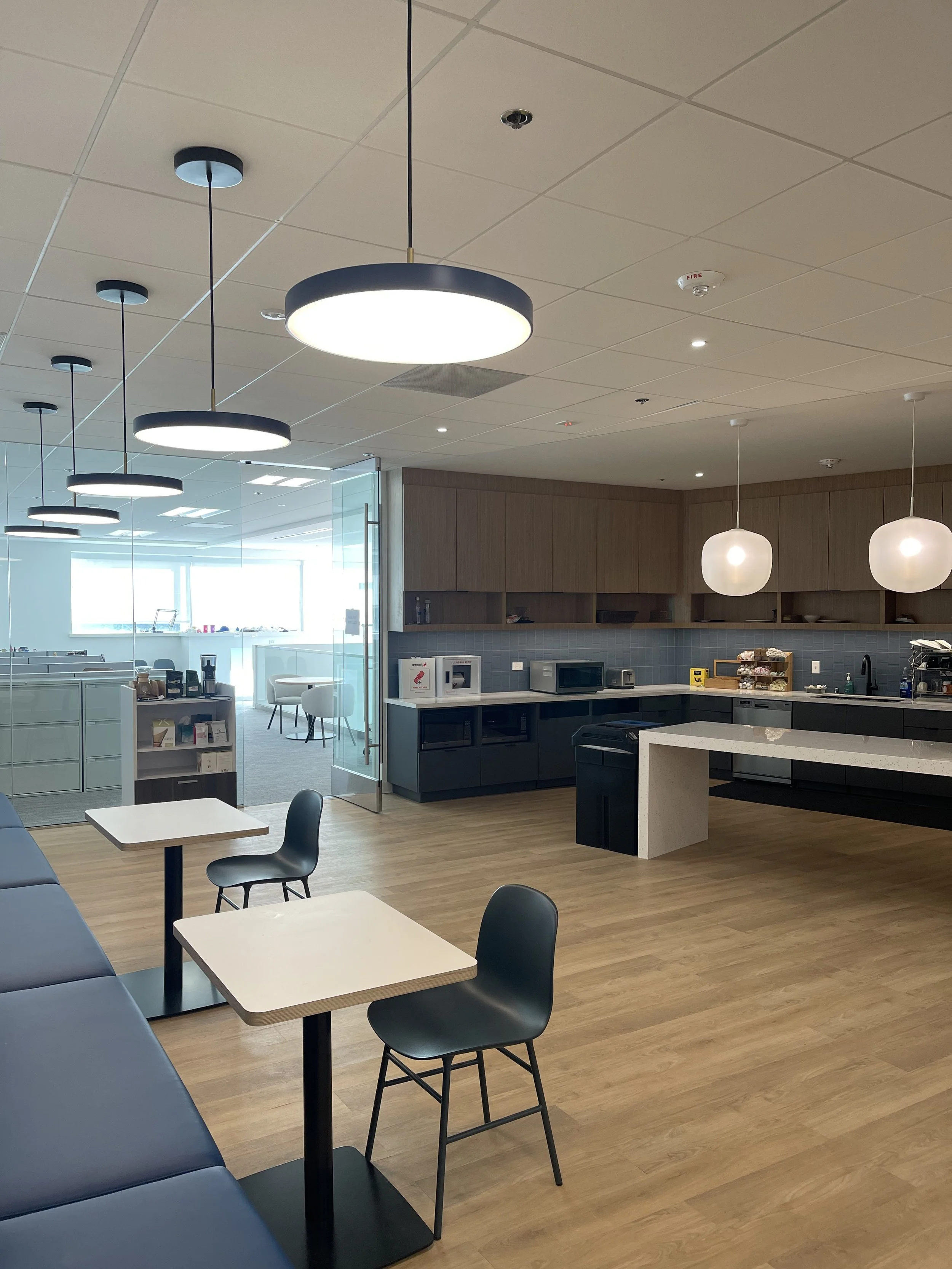 Modern office kitchenette with wooden cabinets, white countertops, and seating area with black chairs and small tables, illuminated by overhead lighting.