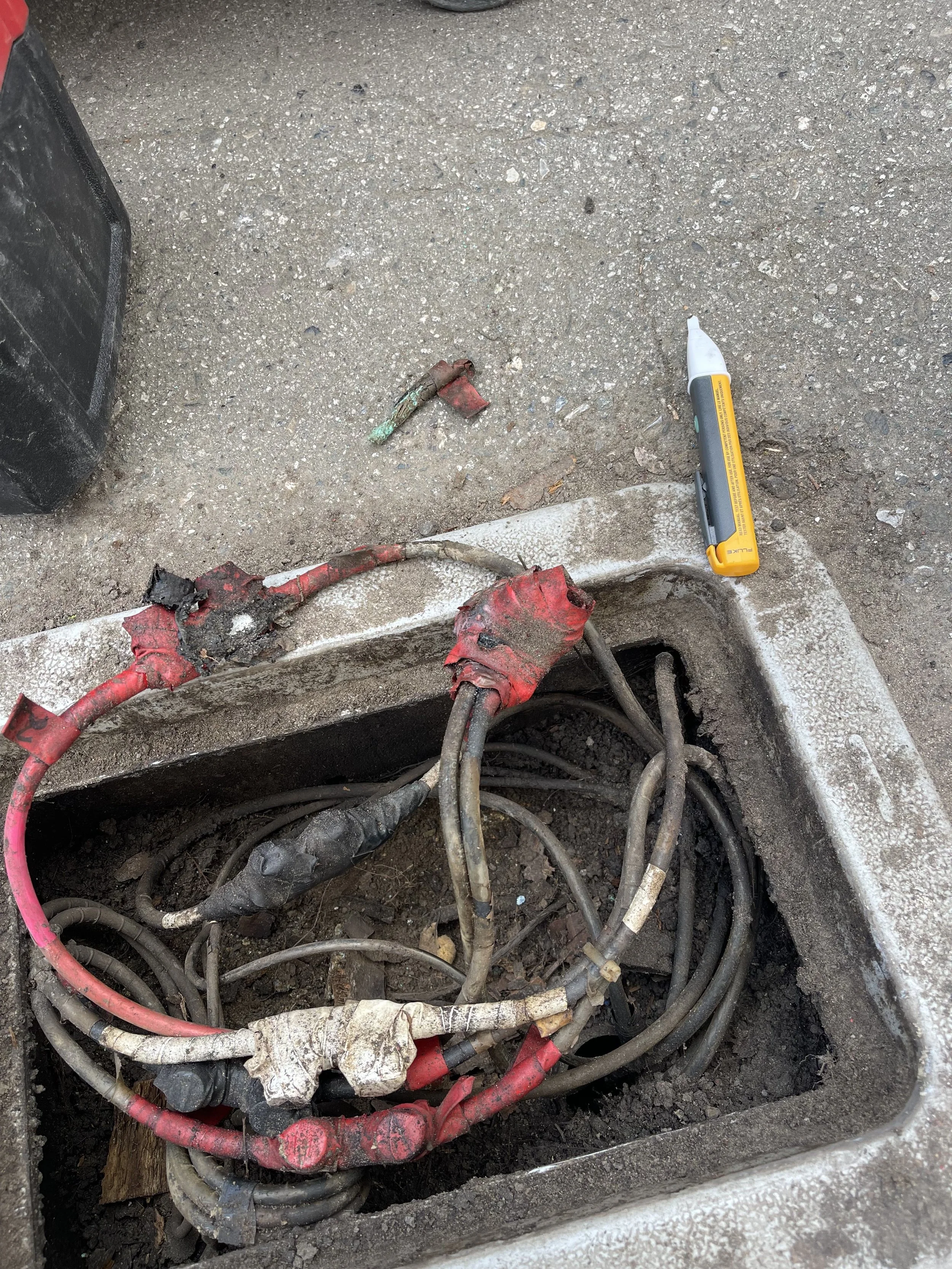 Open electrical box containing multiple damaged and worn electrical wires with some exposed and frayed, on the ground outside. A yellow marker pen and debris next to the box.