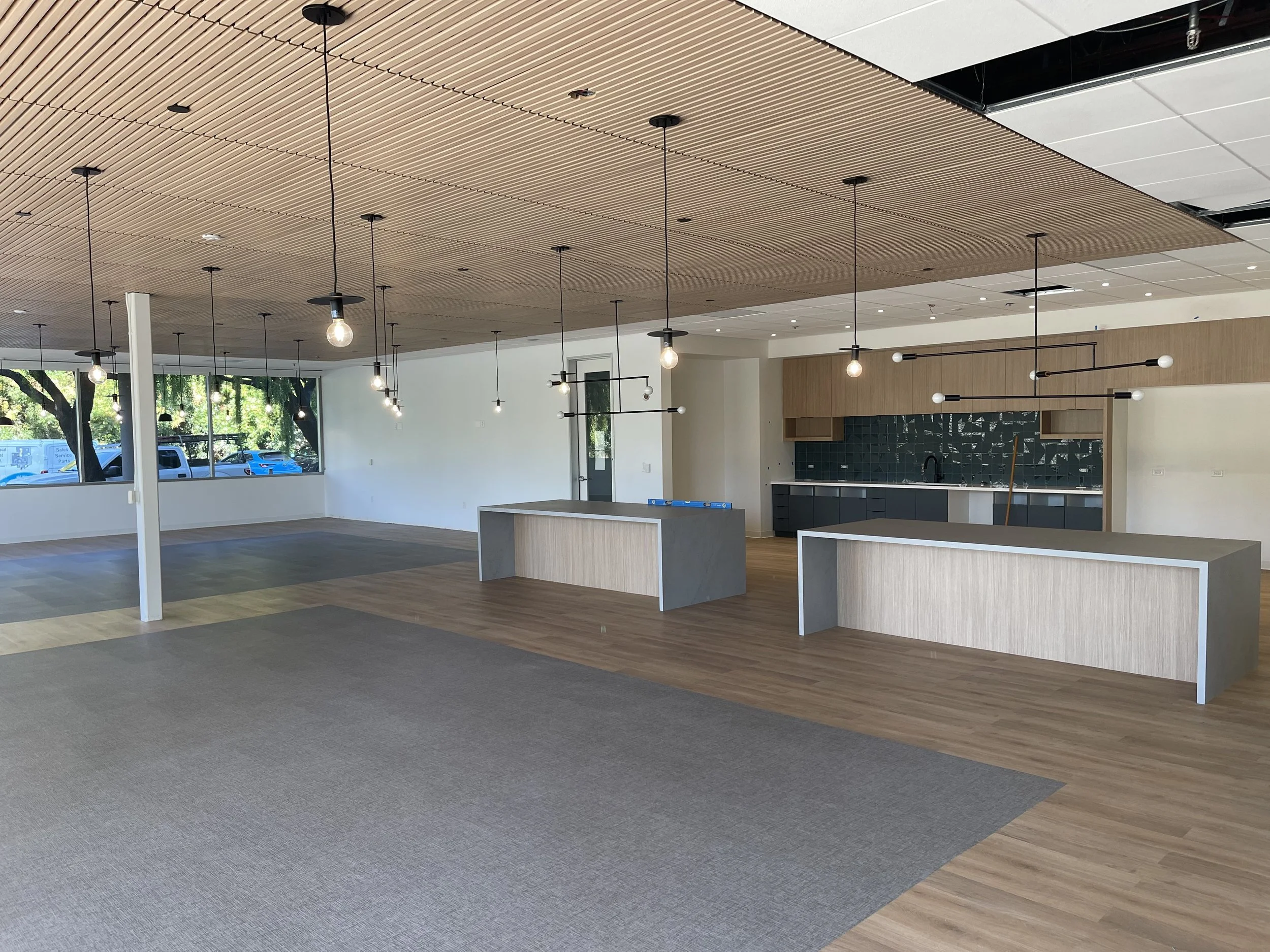 Empty modern interior space with wooden ceiling, pendant lights, large windows, and two kitchen islands with dark cabinets and a teal tile backsplash.
