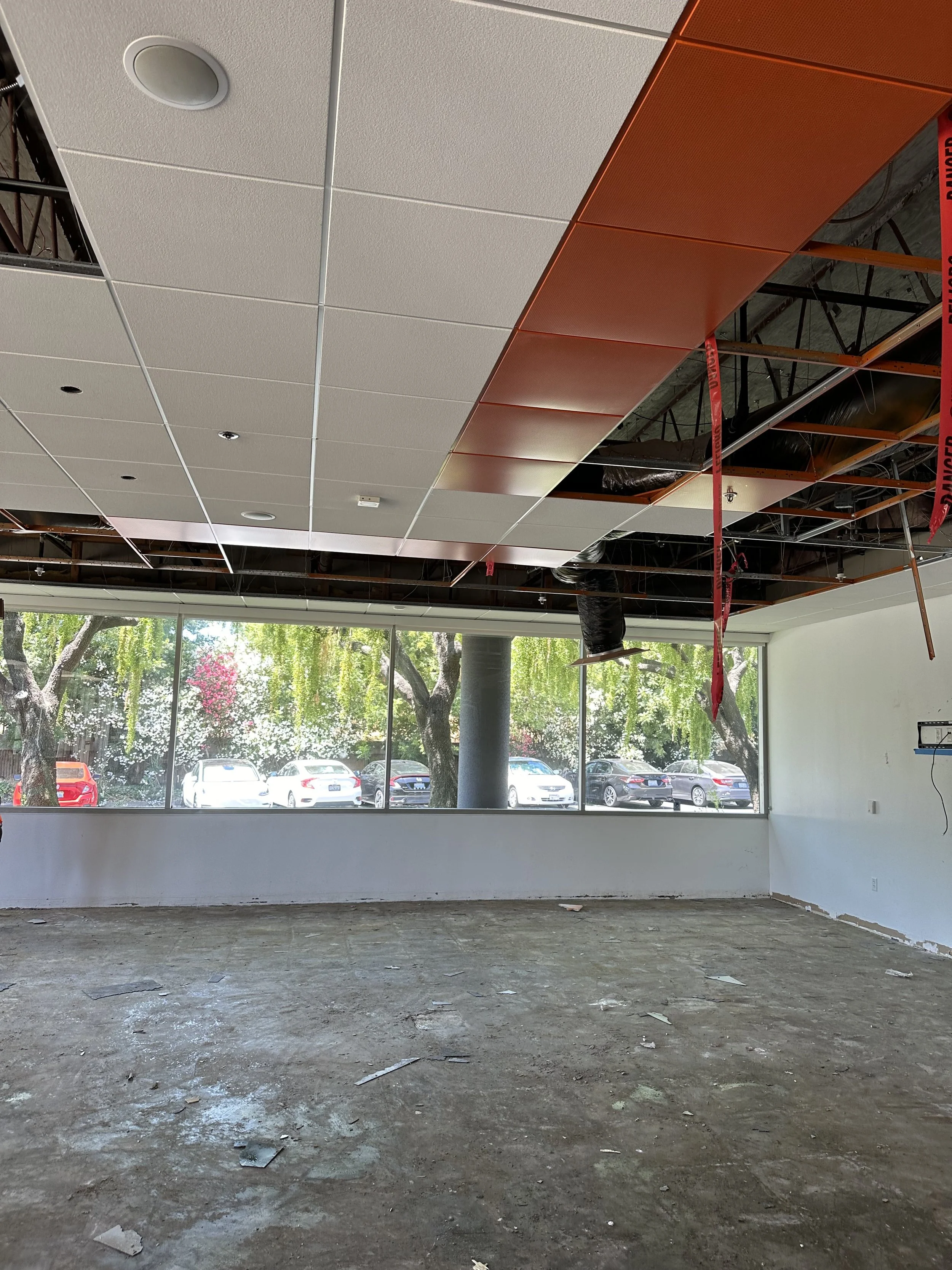 Interior of a room under construction with a suspended ceiling partially installed, exposed ductwork, and a large window showing parked cars and trees outside.