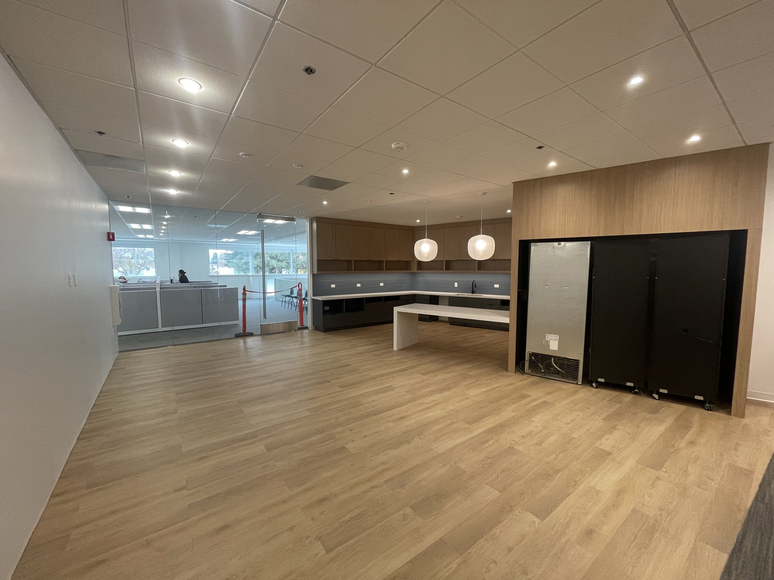 Empty office lounge with wood flooring, modern lighting, a bar-style counter, and a wood-paneled wall with black equipment stored against it.