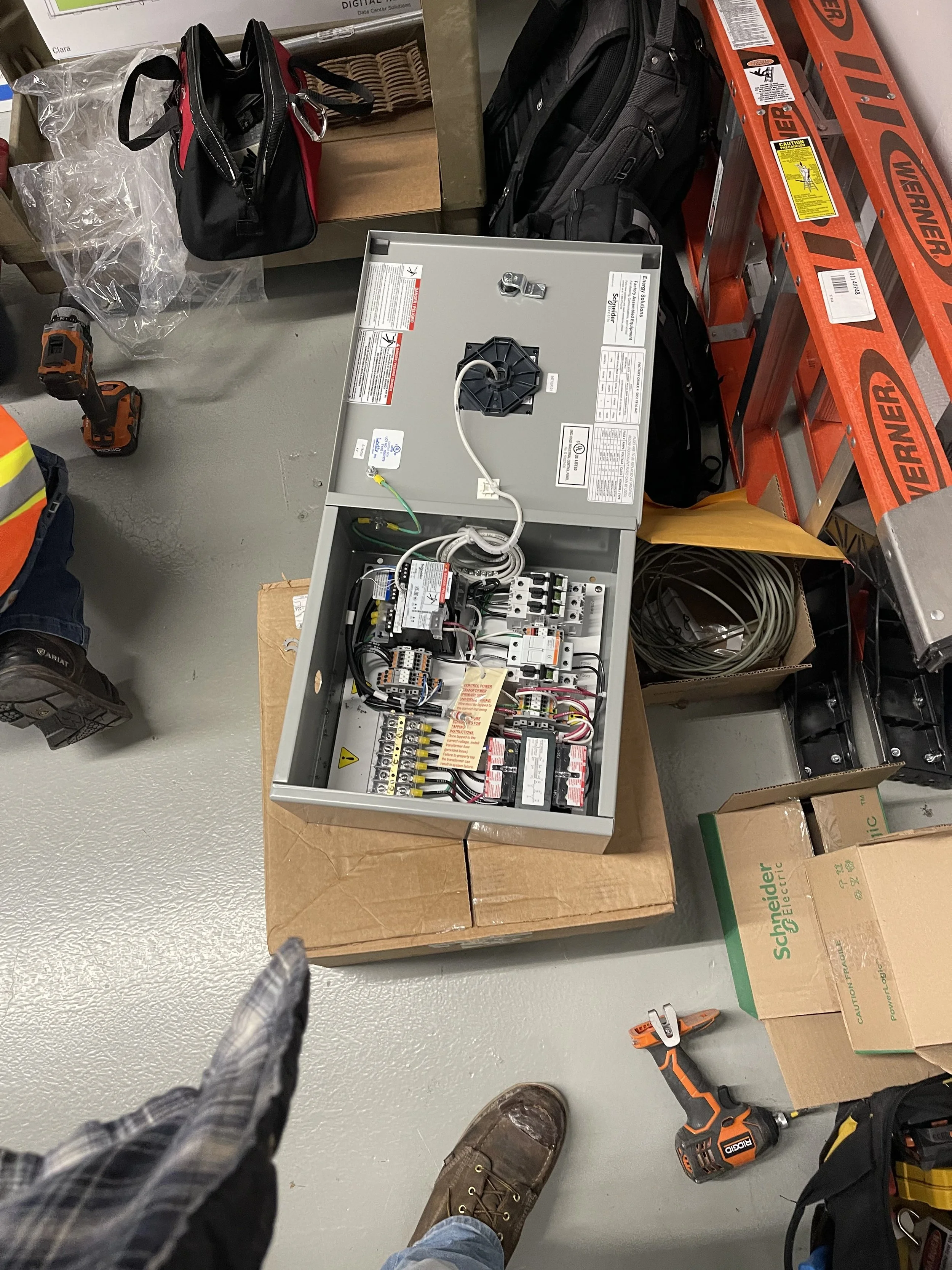 Open electrical control panel with wiring and circuit breakers, placed on a cardboard box in a workshop or utility room. Surrounding items include tools, a black backpack, cardboard boxes, and a red ladder.
