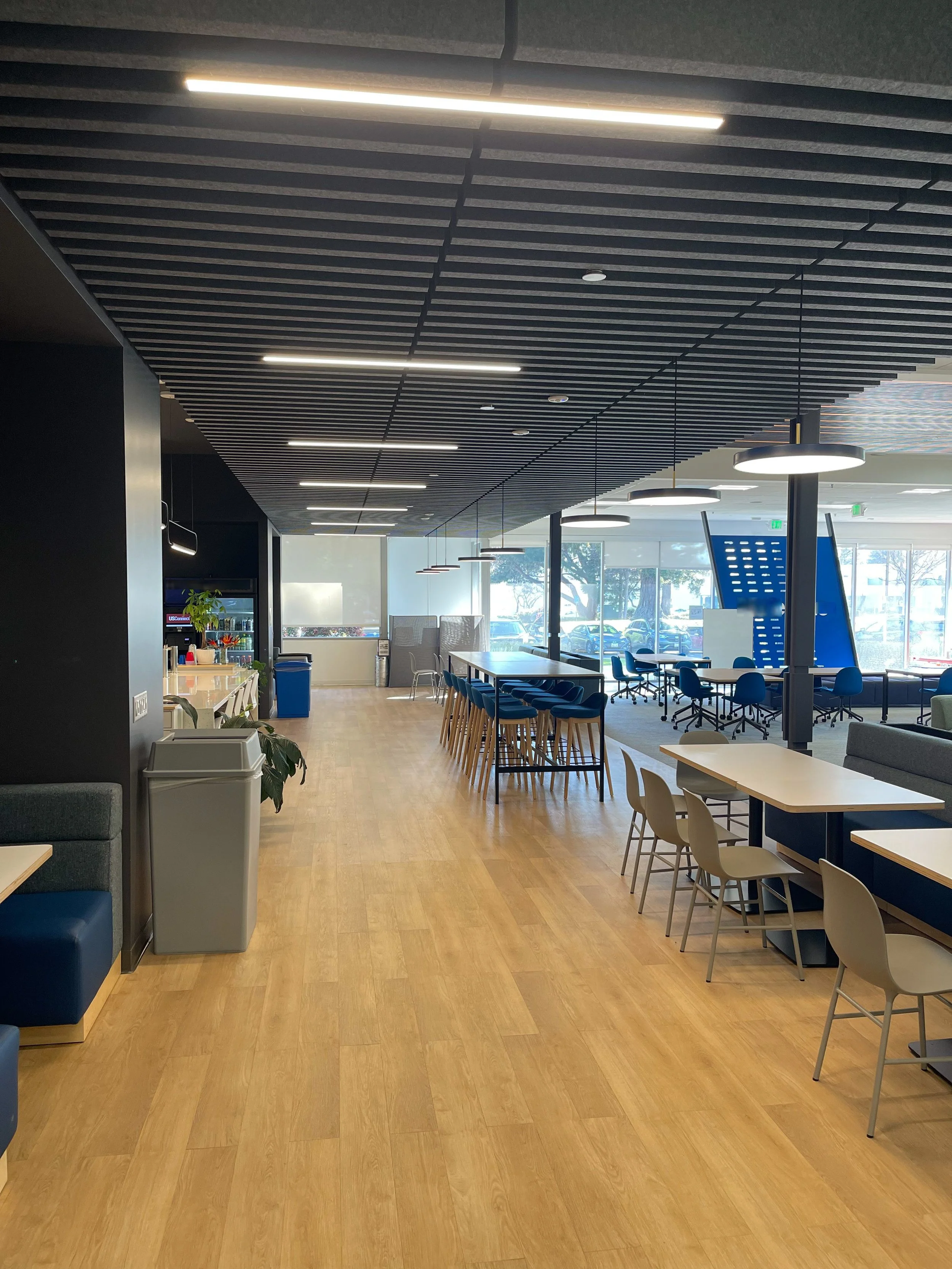 Modern, empty co-working or lounge area with wooden floors, tables, chairs, and large windows letting in natural light.