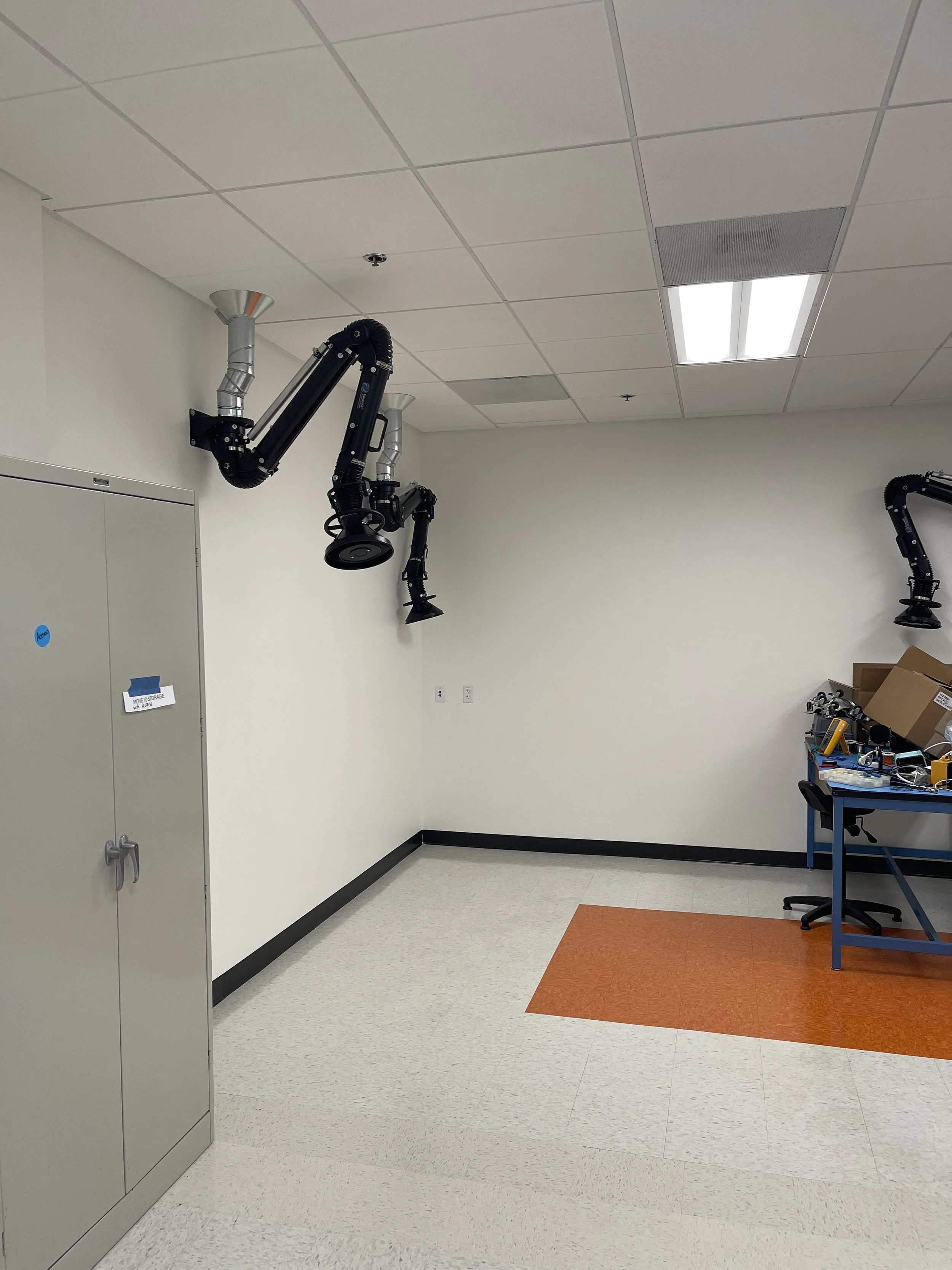 Empty laboratory room with robotic arms mounted on the ceiling, a gray storage cabinet on the left, and a blue work table with tools and boxes on the right