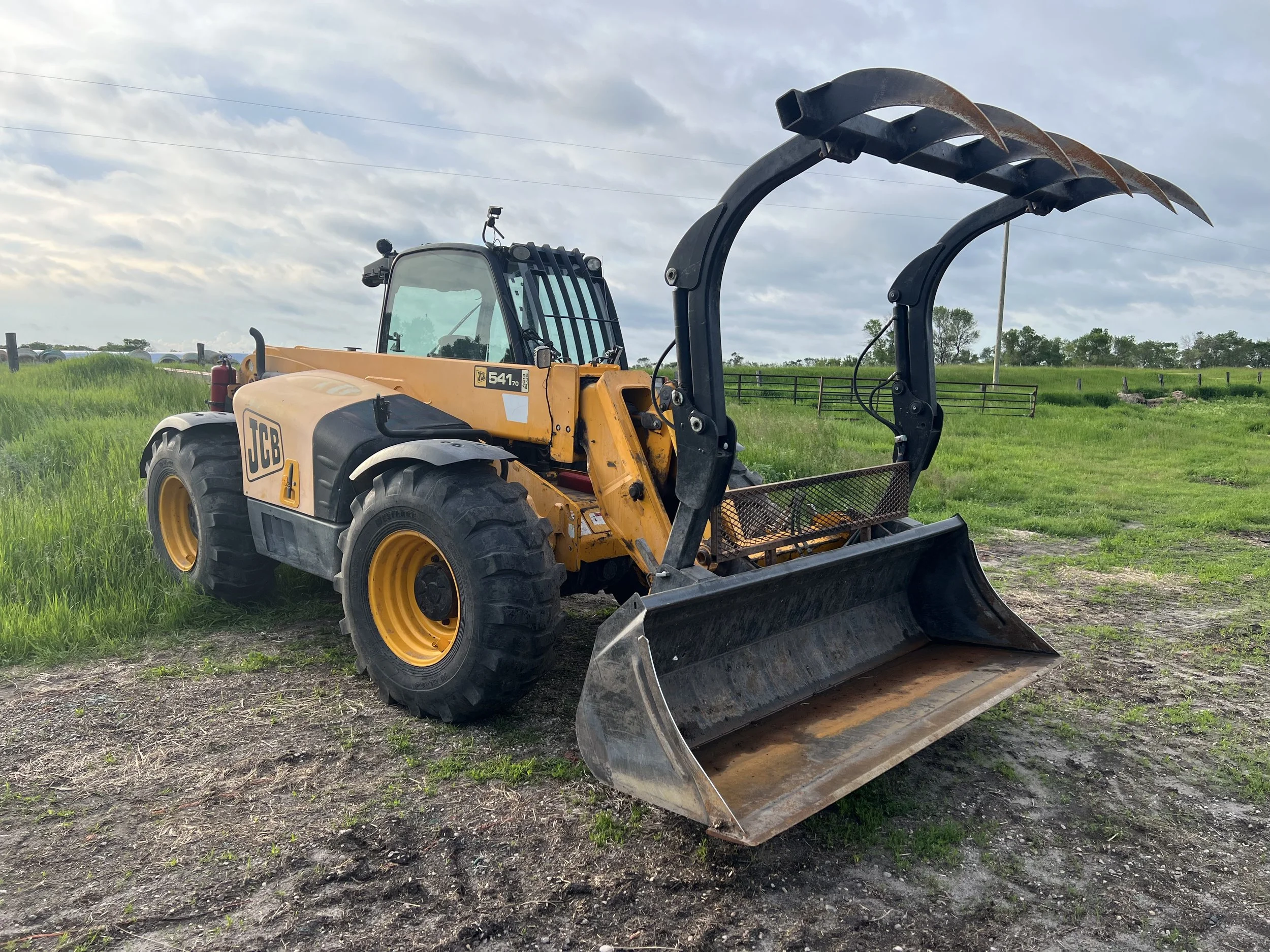 A yellow JCB telehandler with a large metal bucket and grapple attachment ready to load manure, sitting on some nice grass and dirt.