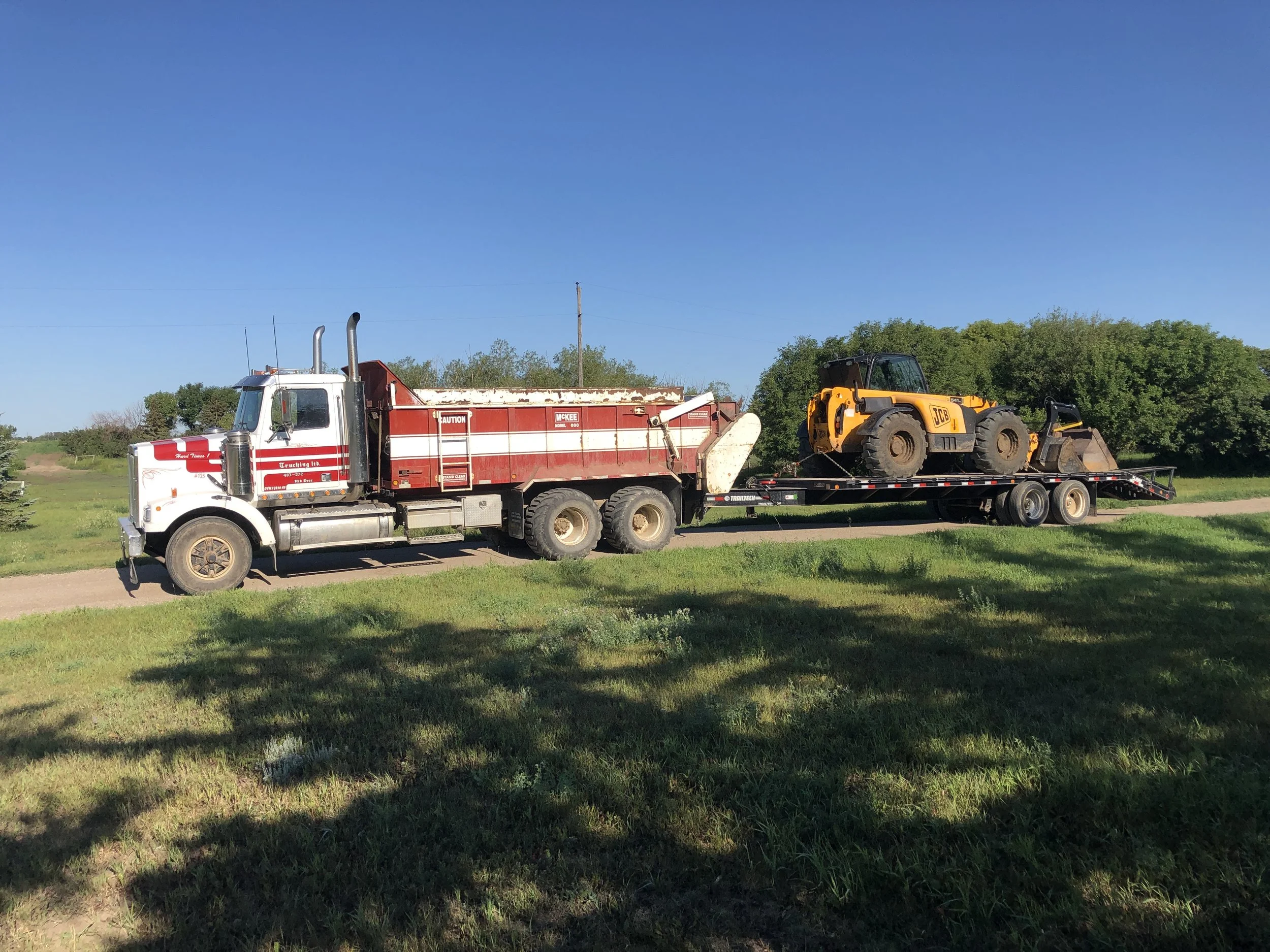 A flatbed truck transporting a yellow JCB telescopic loader on a rural dirt road with green grass, trees, and a blue sky in the background.