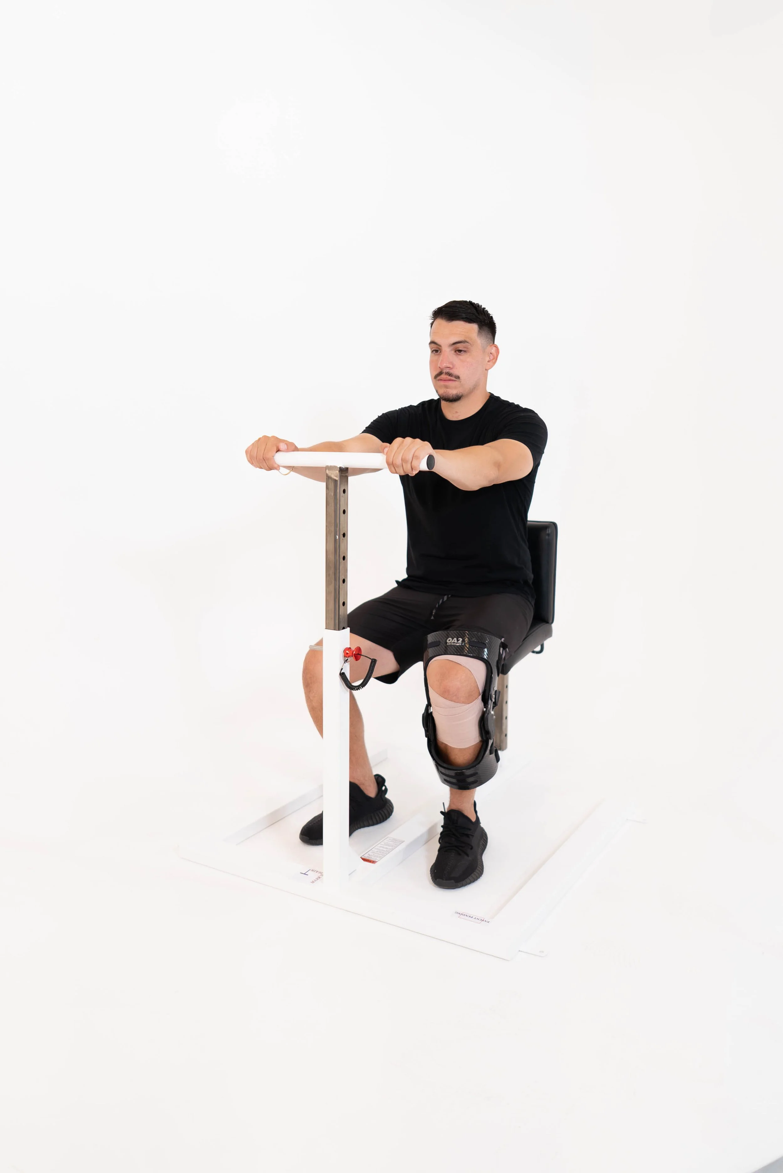 A man sitting on a chair with a knee brace and using a motorized rehabilitation device for leg therapy, in a bright white studio.