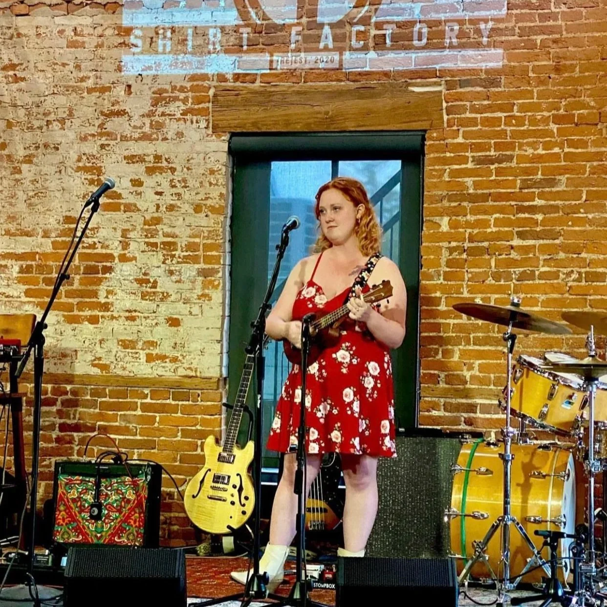 A woman with red hair wearing a red floral dress is standing on stage with a ukulele, surrounded by musical instruments including a guitar and drums, in front of a brick wall with a sign that reads 'The Kit Factory'.