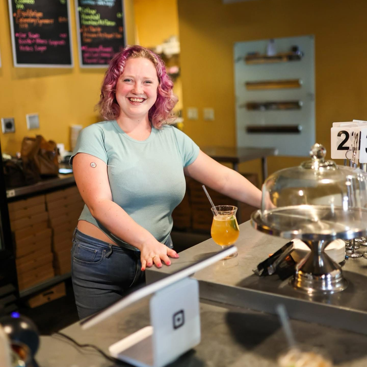 A woman with pink curly hair, smiling, standing behind a counter in a cafe or restaurant, with a beverage in front of her and a menu board in the background.