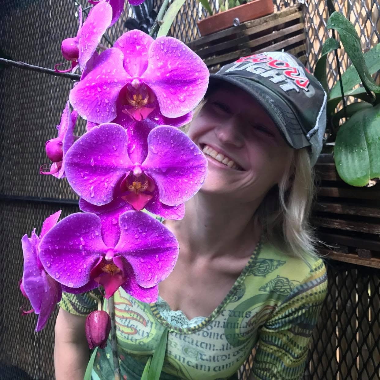 Woman smiling behind a large, vibrant purple orchid flower with water droplets, wearing a baseball cap and a colorful shirt.