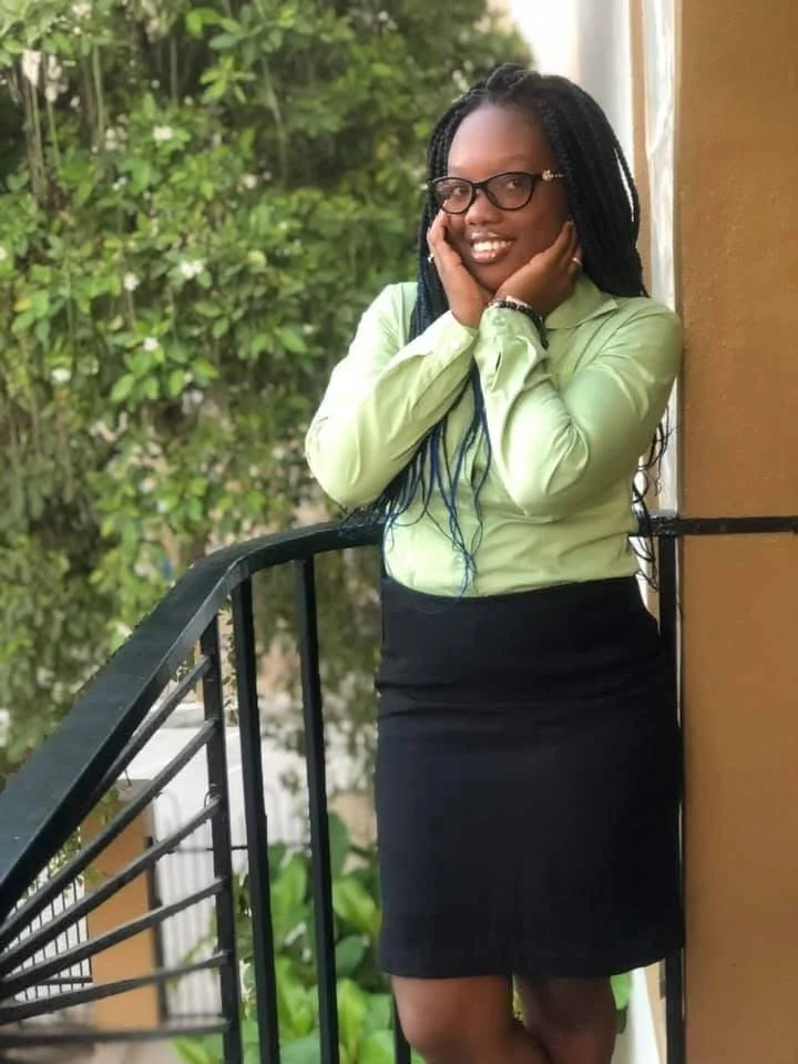 A woman with long black braided hair, wearing glasses, a light green blouse, and a black skirt, is leaning on a black railing on a balcony, smiling and touching her cheeks with both hands, with greenery in the background.