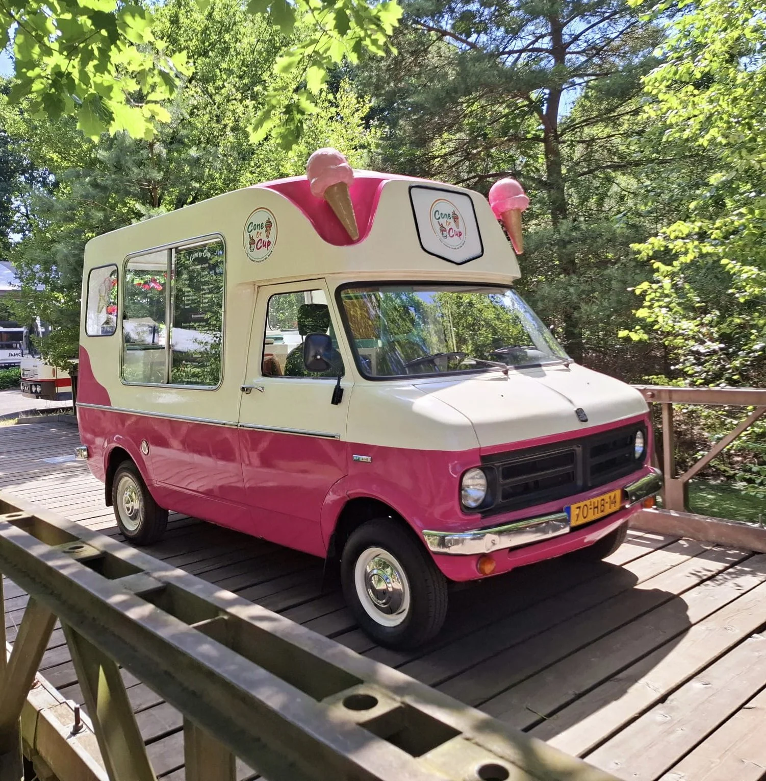 Een kleine ijswagen in roze en crème met ijsco-onderdelen op het dak en logo's met de tekst 'Cone or Cup', geparkeerd op een houten platform in een bosrijke omgeving.