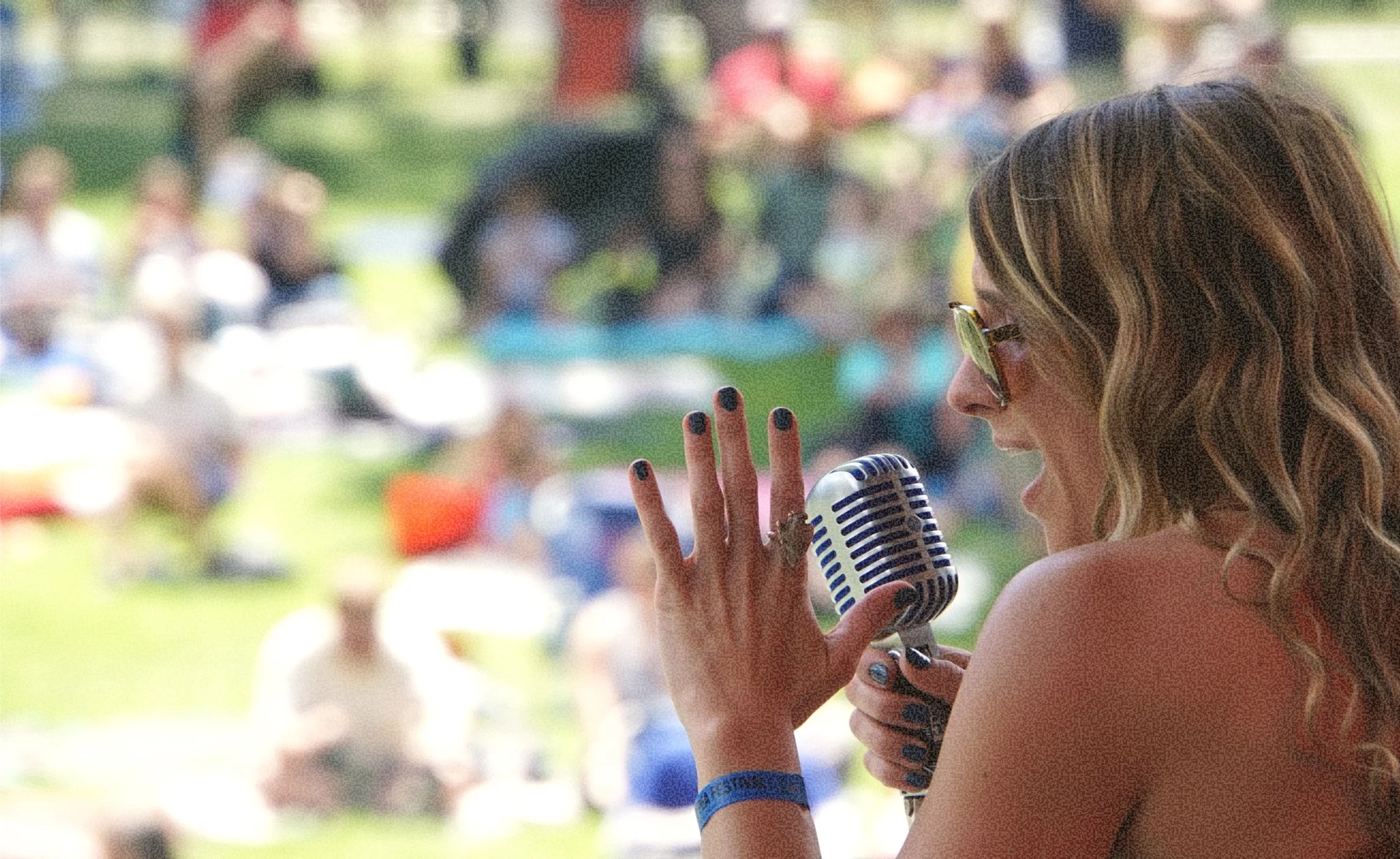 A woman singing into a vintage microphone outdoors, with a blurred festival crowd behind her.
