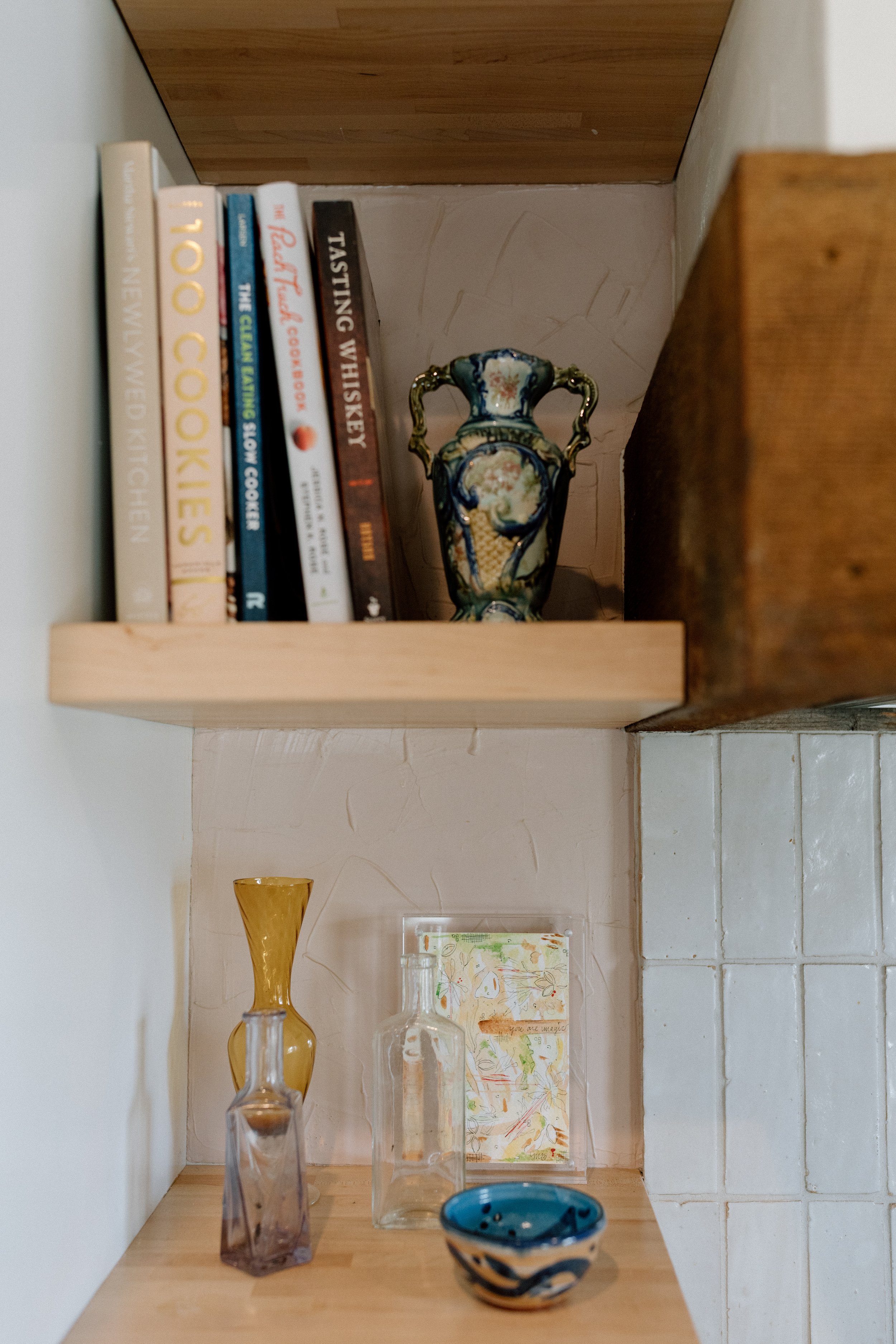 A corner shelf with cookbooks, a decorative ceramic vase, glass bottles, a small decorative bowl, and a colorful postcard.