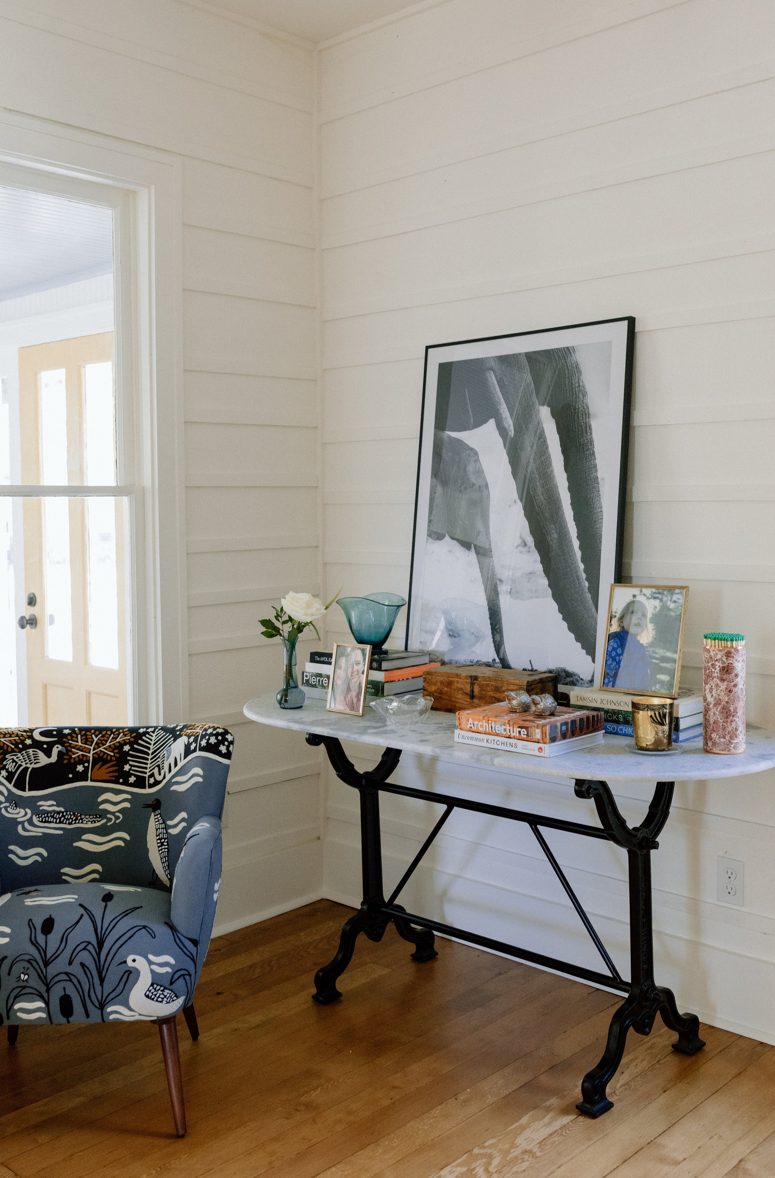 Interior scene with a white wall, a light-colored dresser with decorative items, framed photographs, and large black-and-white artwork leaning against the wall, next to a window with natural light.