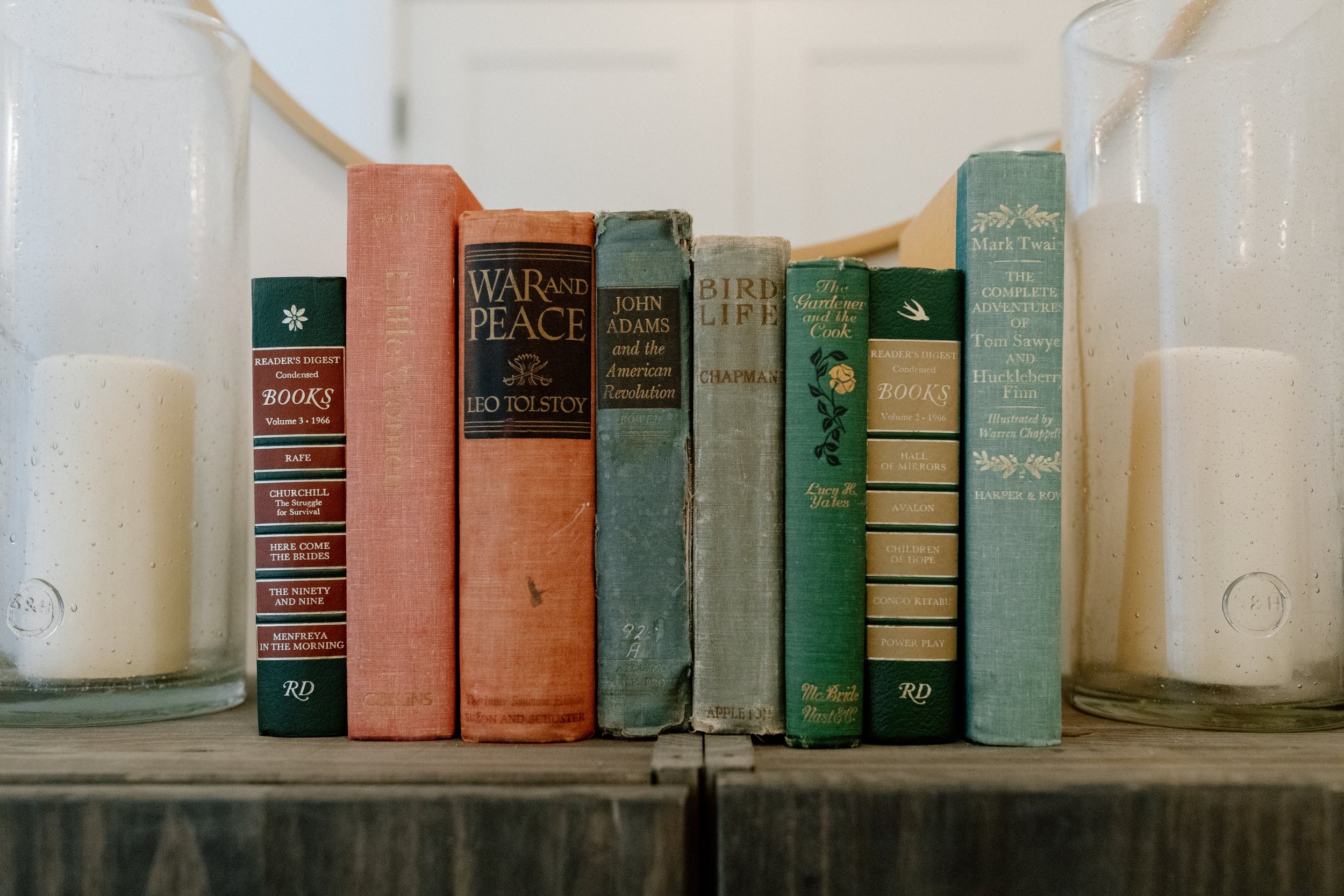 A row of vintage books on a wooden shelf between two glass candle holders with white candles, with a white wall background.