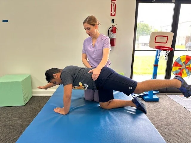 A woman appears to be a physical therapist or trainer assisting a man with a back bend exercise on a blue mat in a rehab or therapy room with sports equipment and a fire extinguisher in the background.
