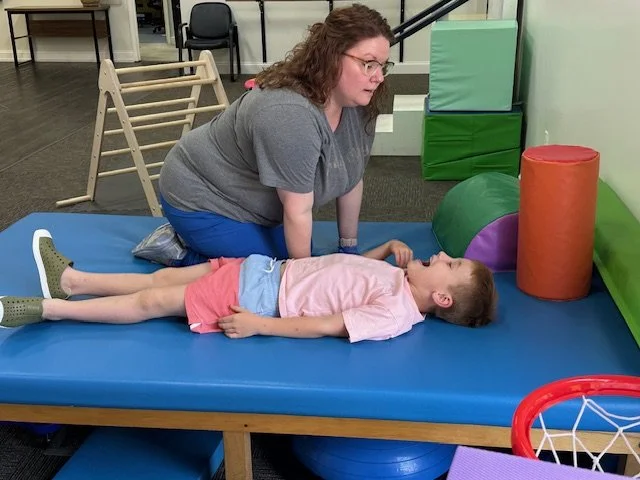 A woman assists a child lying on a therapy mat in a therapy room with padded walls and colorful foam shapes.