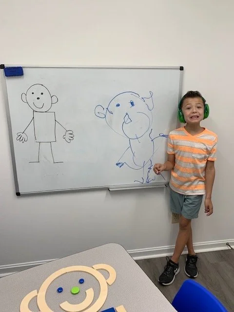 A young boy standing next to a whiteboard with two drawings of cartoon characters, one simple stick figure and one large-headed, expressive character, in a room with a gray floor and a small table with a teddy bear-shaped object and colorful items.