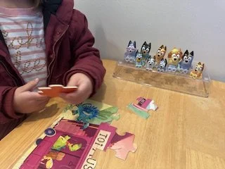 Child in maroon jacket playing with a card game on a wooden table, with a clear case of small, colorful animal figurines in the background.