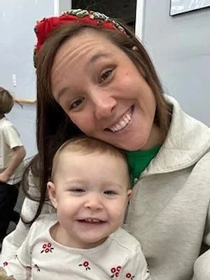 A woman smiling happily with a young child, both looking at the camera, indoors with other people in the background.