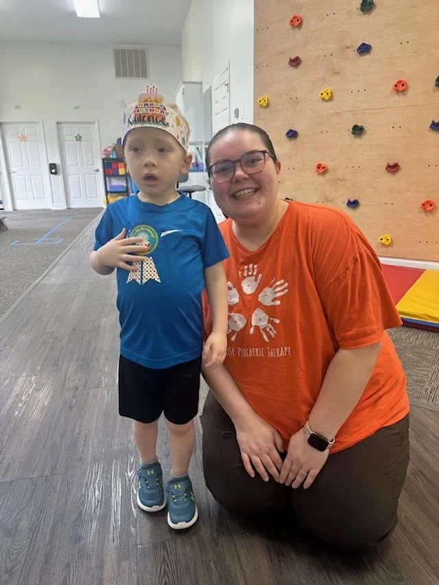 A young boy wearing a birthday hat and a woman kneeling next to him in an indoor play area or therapy center, with a climbing wall in the background.
