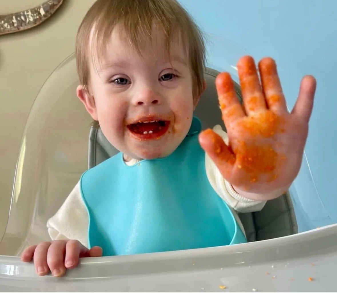 A young child with messy food on their face and hand, sitting in a high chair and showing a smiling, food-covered hand to the camera.
