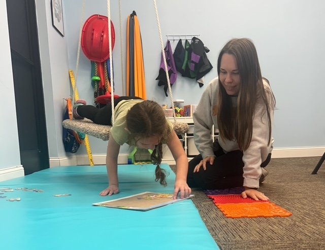 A girl and a woman reading a book together on the floor in a room with colorful sports and outdoor gear hanging on the wall.