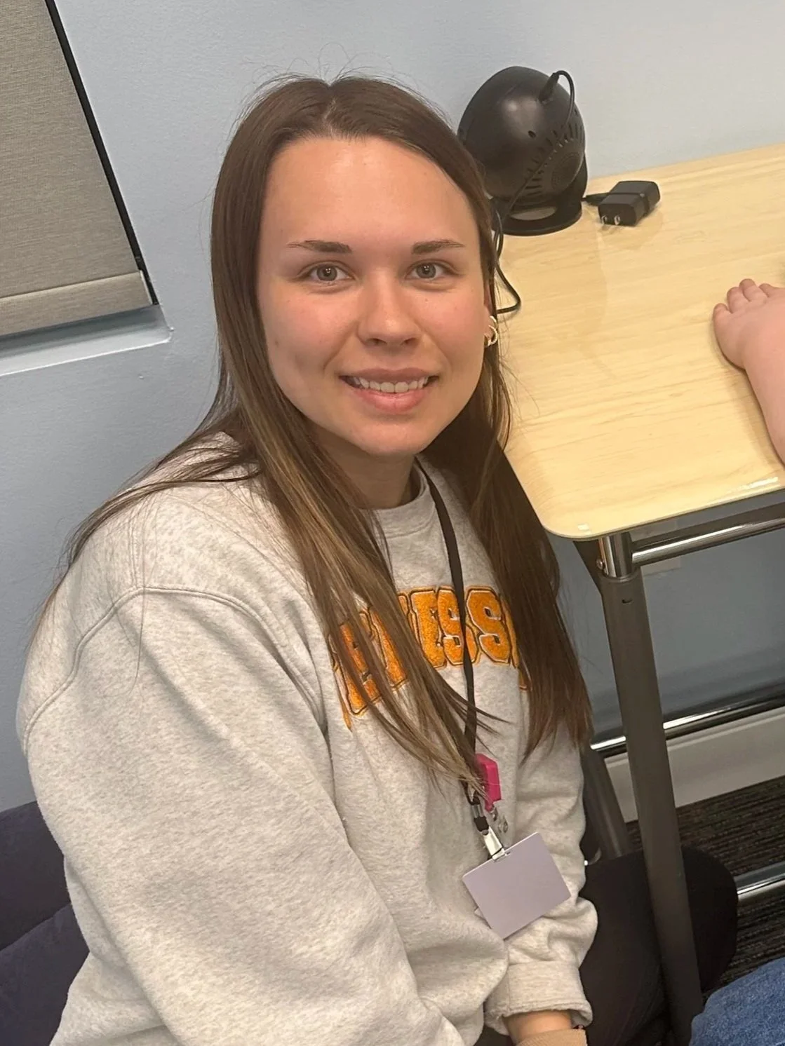 Young woman with long brown hair and a gray sweatshirt, sitting on the floor next to a light wooden table with a black headset on, smiling at the camera.