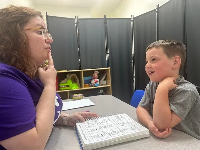 A woman and a young boy sitting at a table, facing each other, engaging in conversation. The woman has curly hair, glasses, and is wearing a purple shirt. The boy has short hair, is smiling, and resting his head on his hand. There are papers and coloring books on the table, and a black folding screen and a shelf with toys in the background.