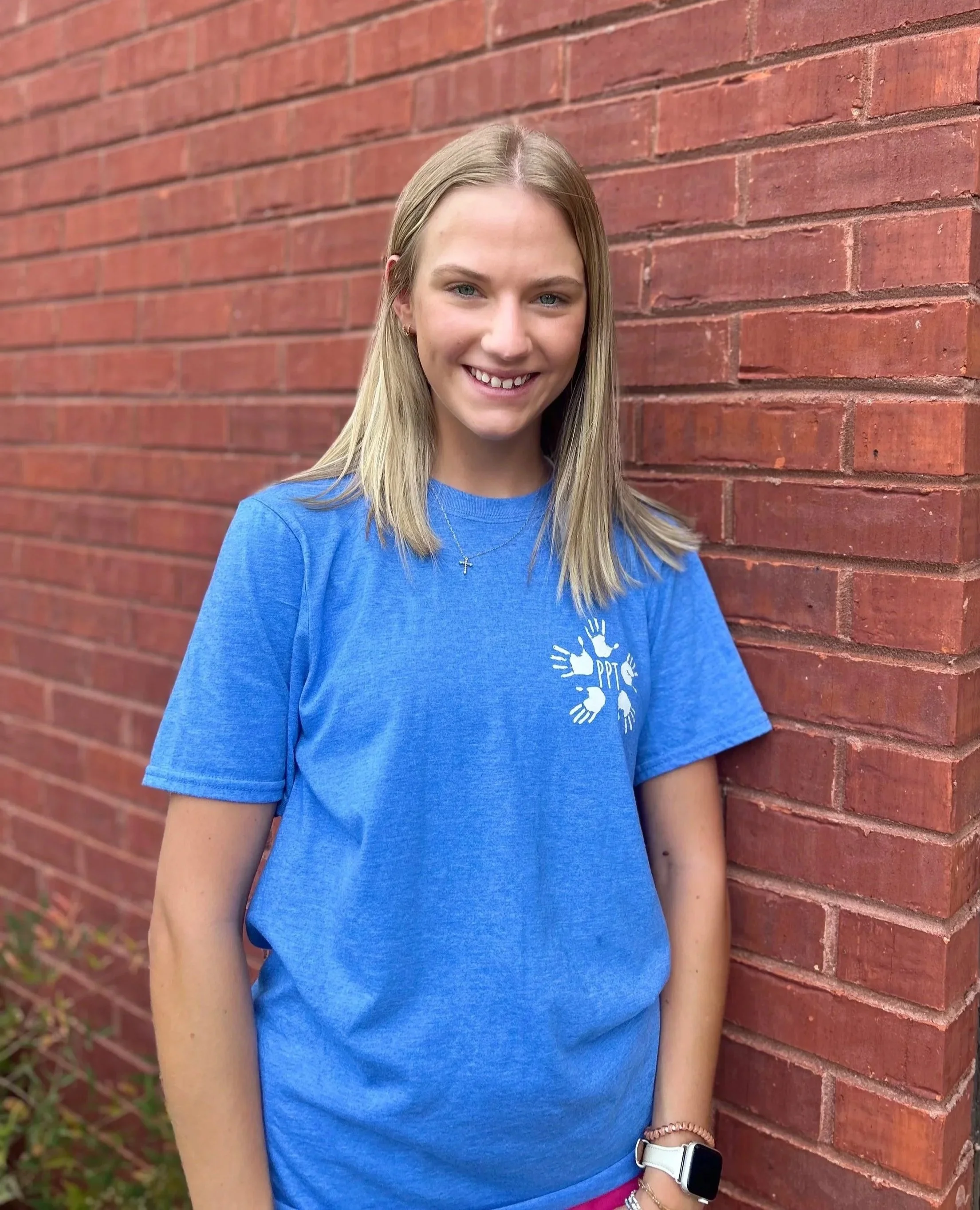 Young woman with blonde hair smiling, wearing a blue T-shirt with a white graphic and a cross necklace, standing outdoors against a brick wall.